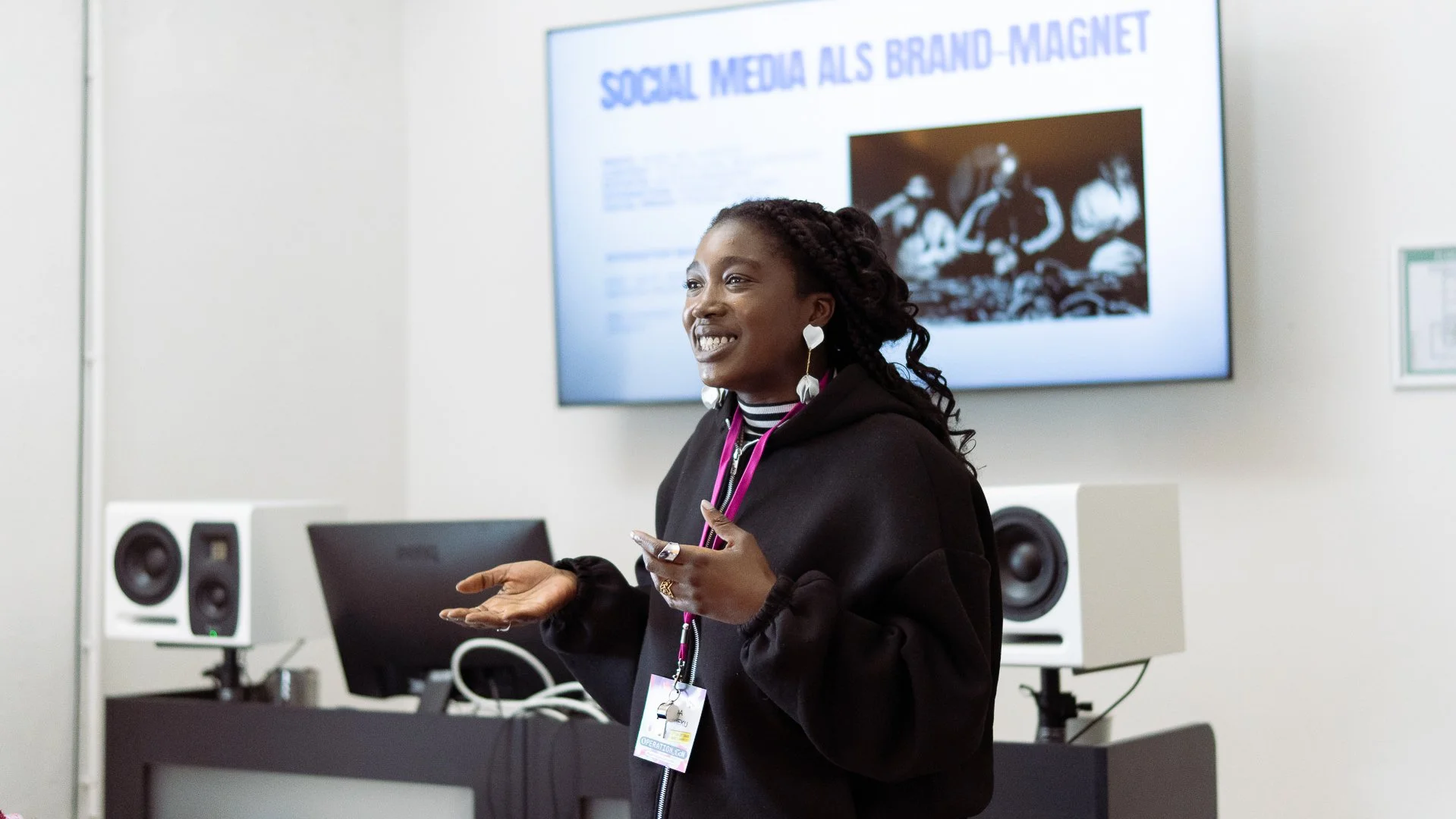 A woman giving a presentation in a conference room, with a screen displaying a slide titled 'Social Media als Brand-Magnet', and speakers visible in the background.
