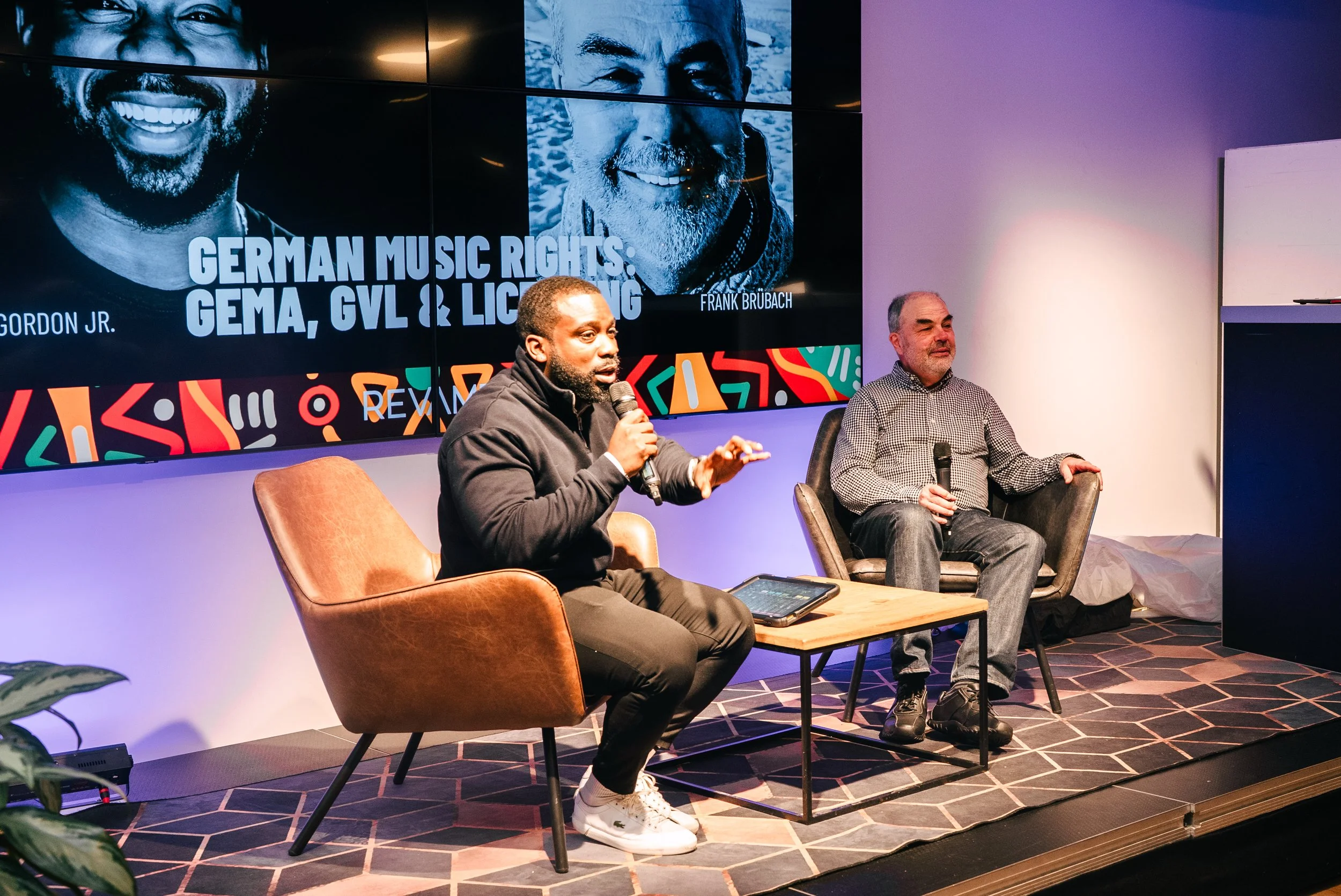 Two men sitting on a stage, participating in a discussion. The man on the left is speaking into a microphone, wearing a black jacket, and sitting in a brown chair. The man on the right is also holding a microphone, dressed in a checked shirt and jean