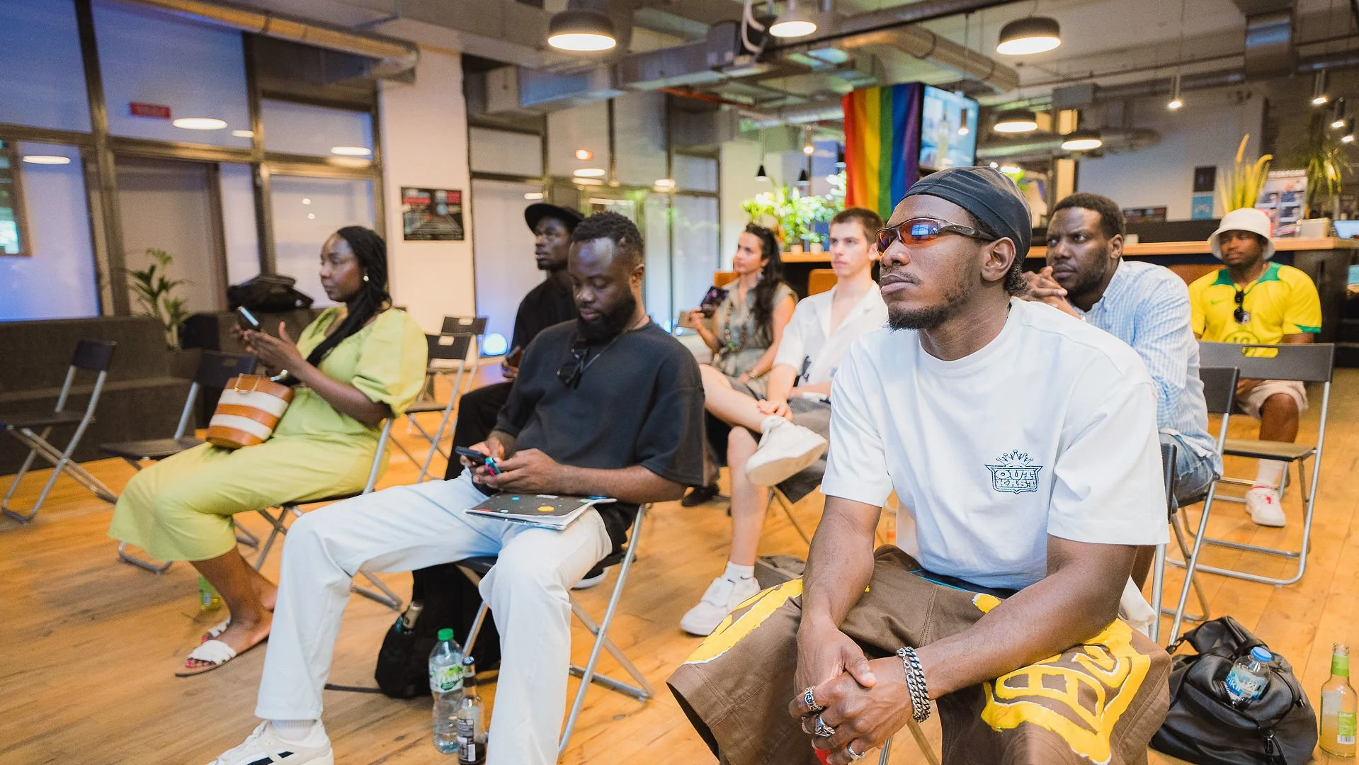 People attending a presentation or seminar in a modern indoor space with concrete walls, wooden floors, and large windows. Some are engaged with their phones, while others are listening attentively.