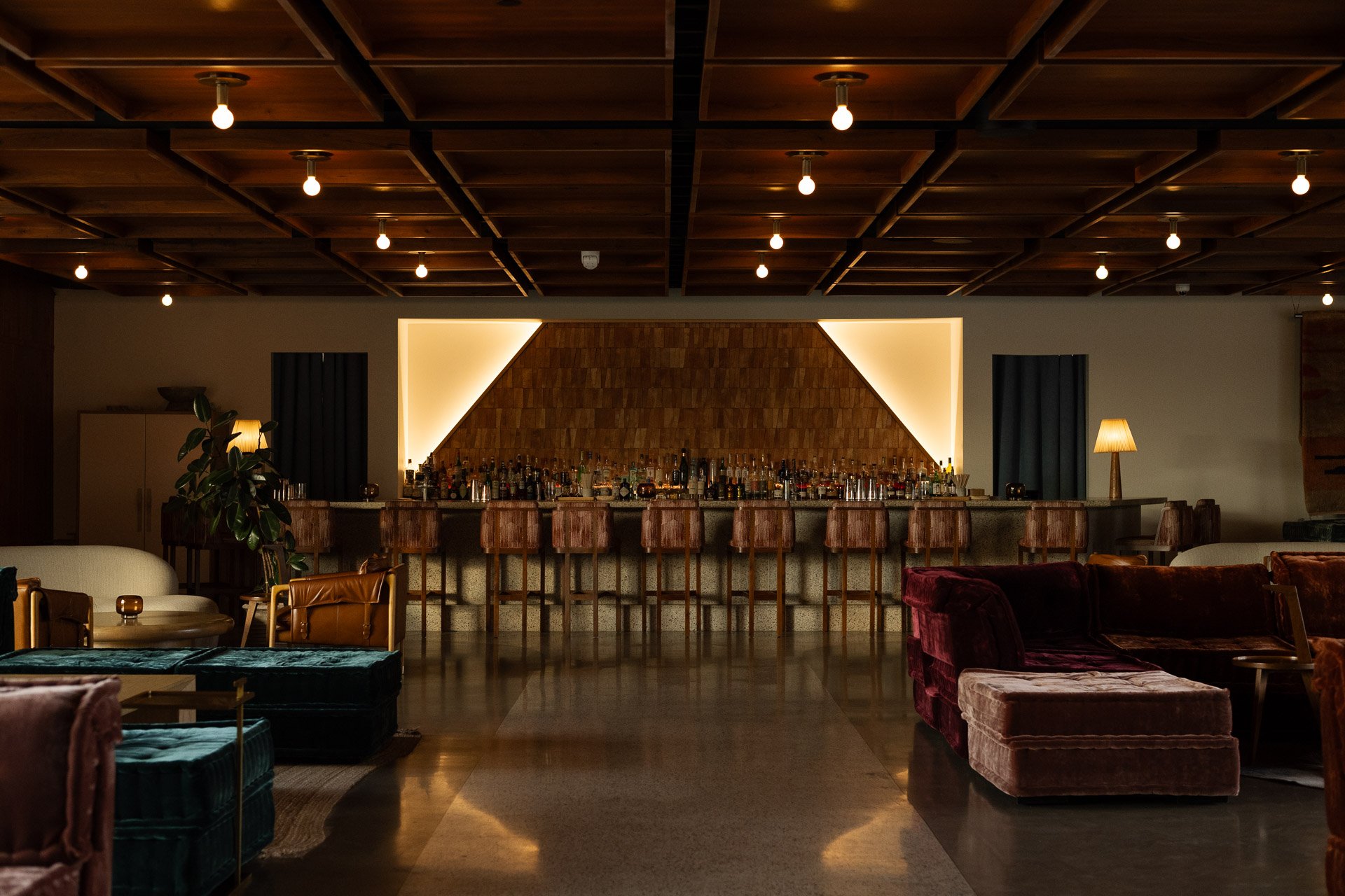 A cozy bar with a long counter lined with bar stools, warm lighting, and plush seating areas, including velvet armchairs and couches in a dimly lit lounge setting.