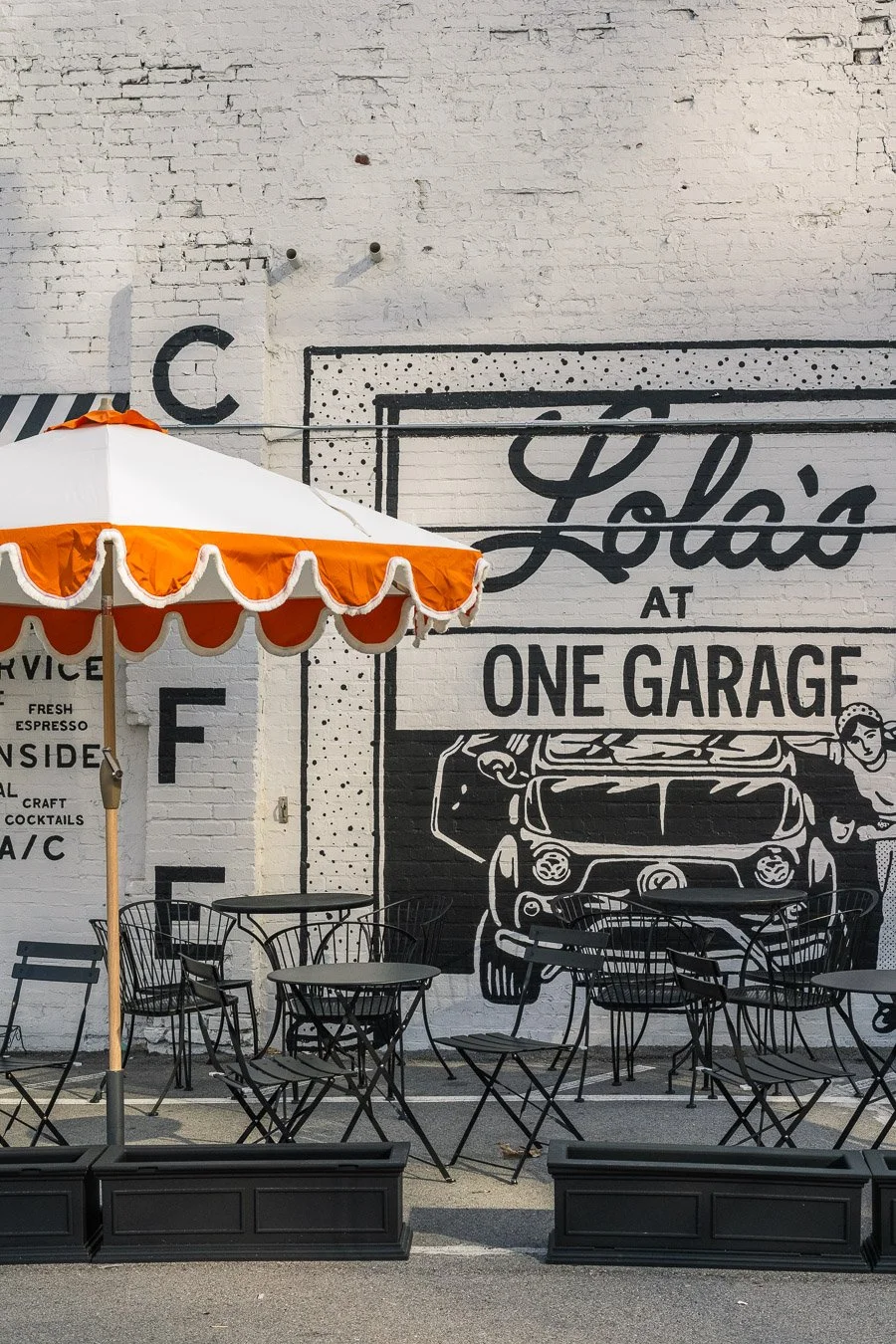 Outdoor seating area with black metal chairs and tables, partly shaded by a large white and orange striped umbrella, in front of a white brick wall with black graffiti-style lettering reading 'Lola's at One Garage' and a drawing of a car.