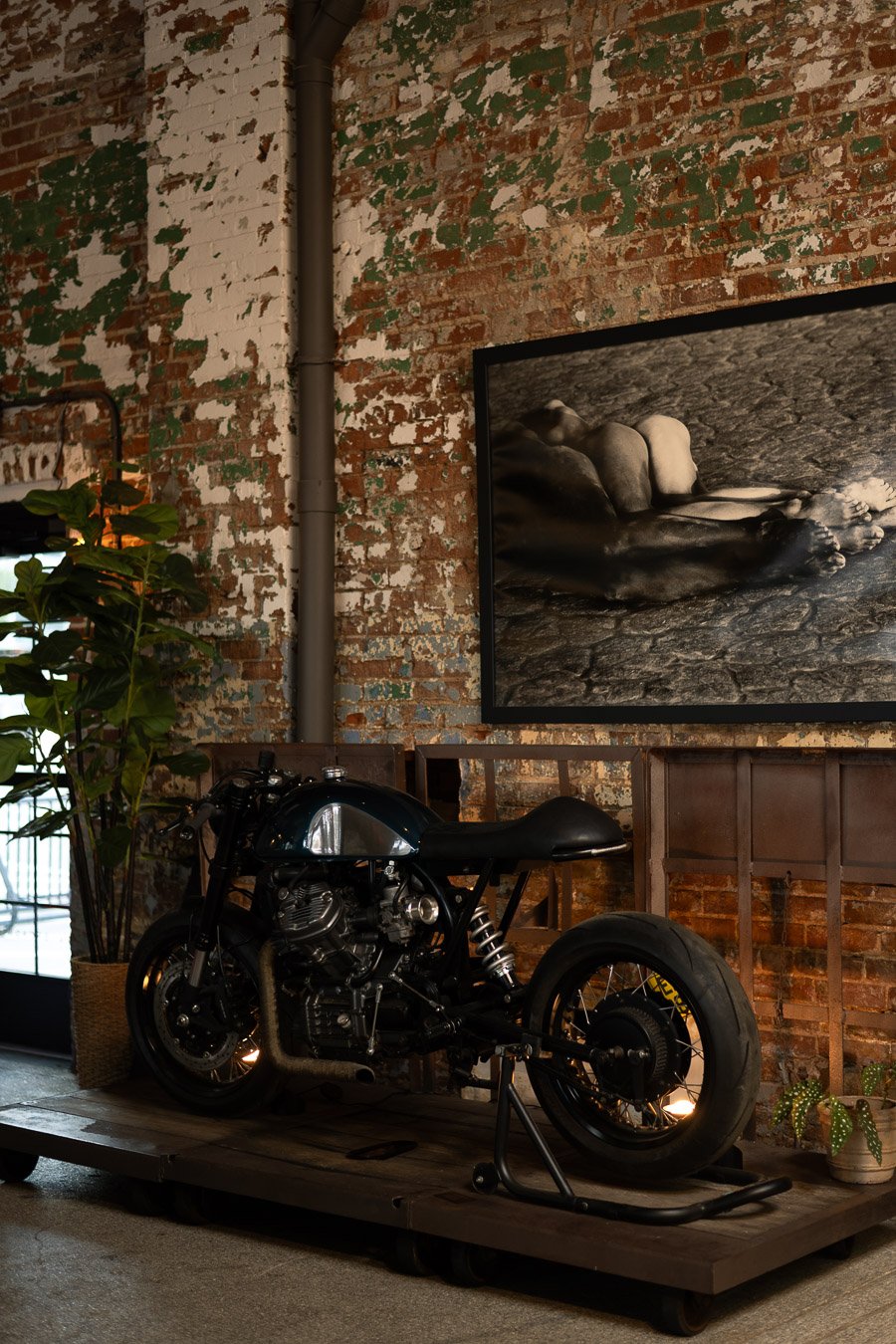 Interior of a rustic industrial space with a vintage black motorcycle on a wooden platform, a brick wall with peeling white and green paint, a framed black-and-white photograph of stones, and potted plants.