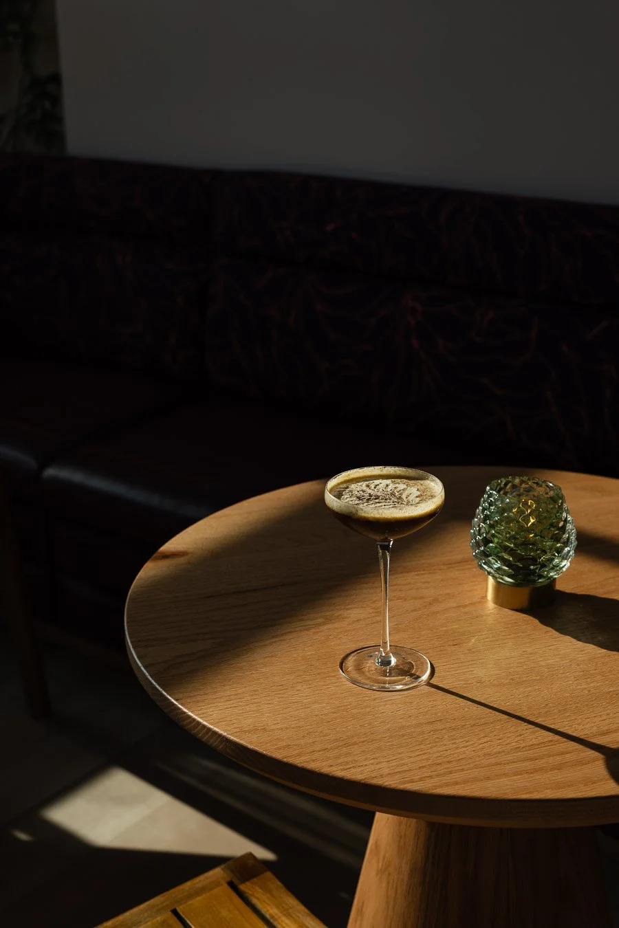 A cocktail glass with a dark drink on a wooden table next to a decorative green glass container, with sunlight casting shadows.