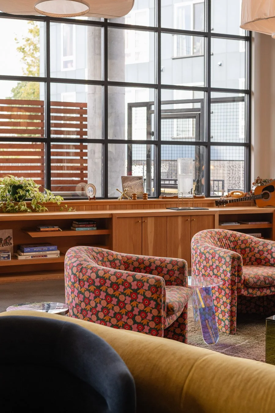 Interior of a cozy room with large window, colorful floral armchairs, a wooden bookshelf, plants, a guitar, and decorative items.