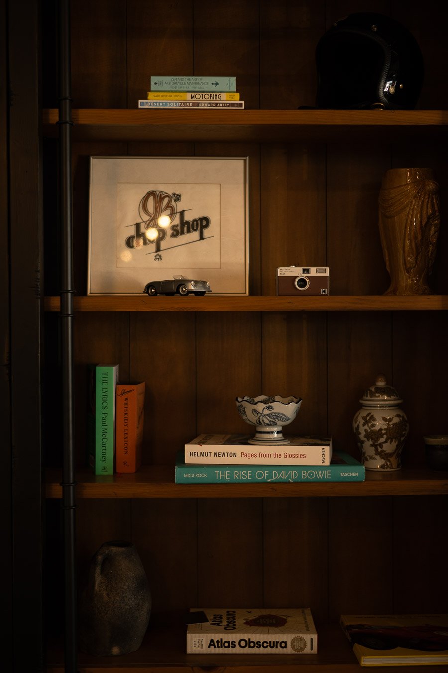 Wooden shelving unit with books, decorative items, and collectibles. Top shelf has books and a black helmet. Middle shelf features a framed print, toy car, camera, and a decorative vase. Bottom shelf holds more books, a ceramic bowl, a ceramic vase, 