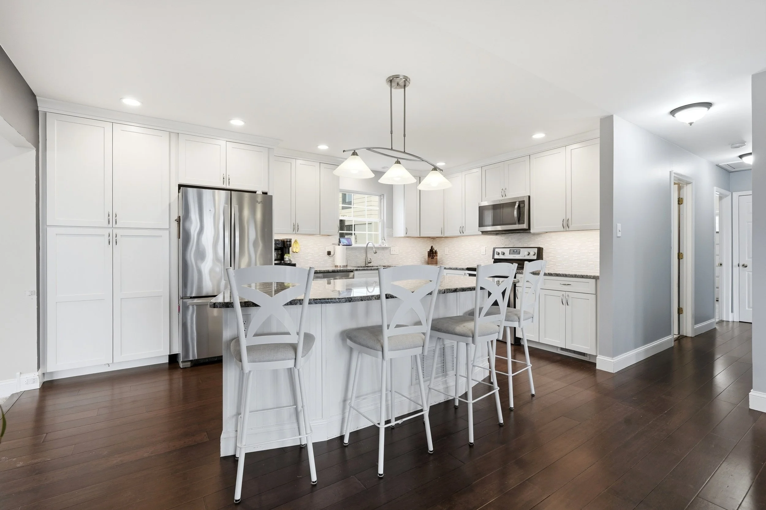 Bright, modern kitchen with white cabinets, stainless steel refrigerator, and a kitchen island with four white chairs. There are multiple ceiling lights and a window in the background.