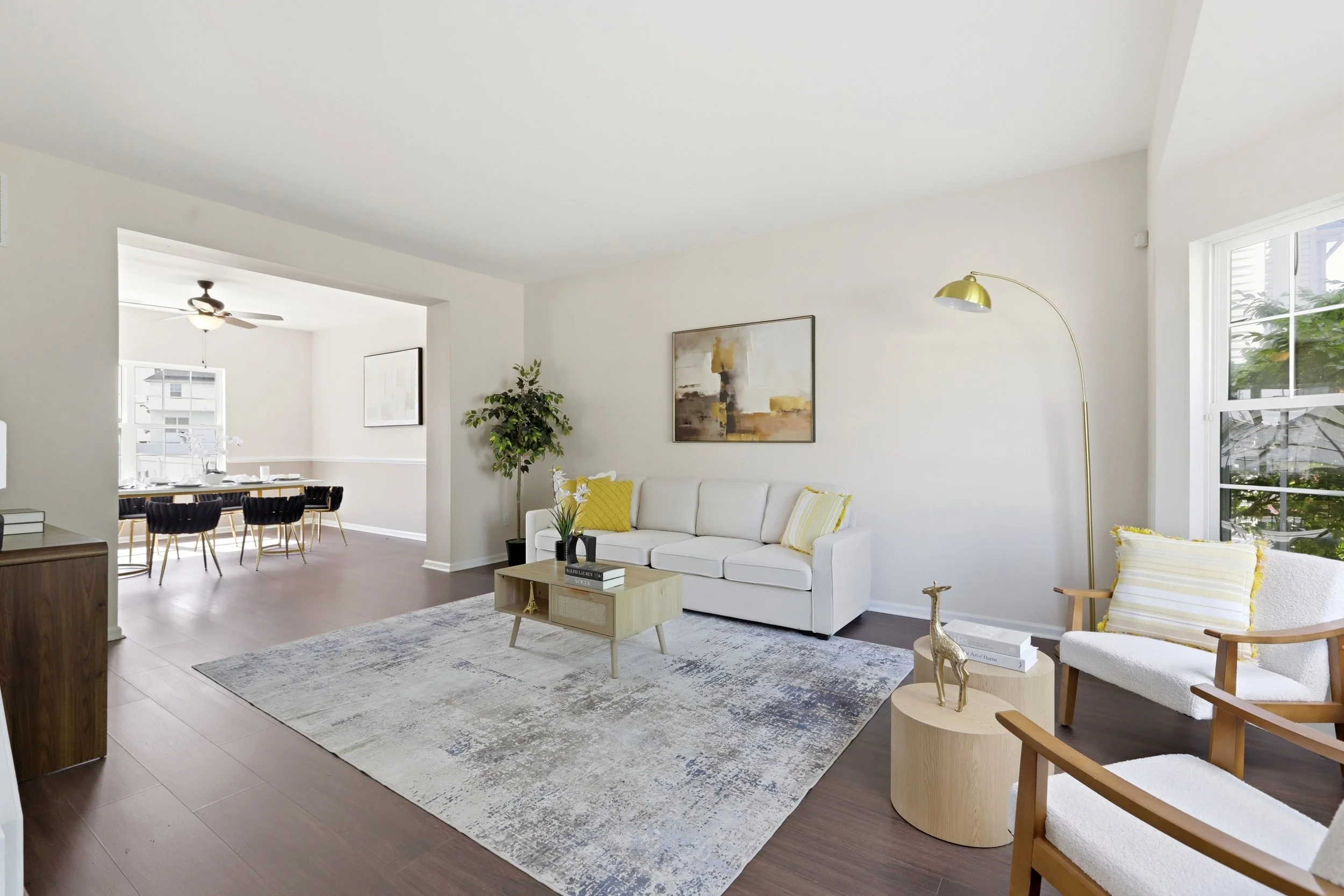Modern living room with white sofa, wooden coffee table, decorative pillows, abstract art on the wall, gold floor lamp, armchair with cushion, and large windows allowing natural light.