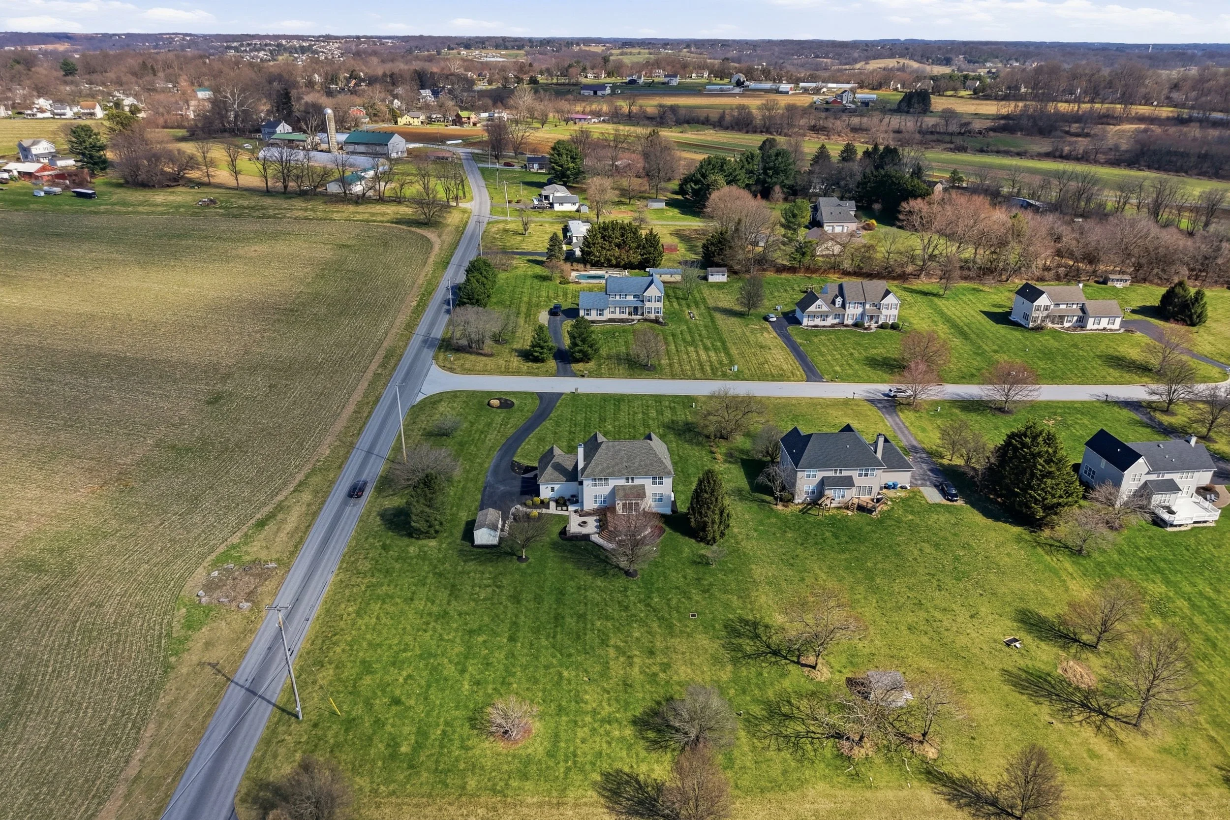 Aerial view of a rural neighborhood with large houses, green lawns, trees, and surrounding farmland, including fields and farm buildings.