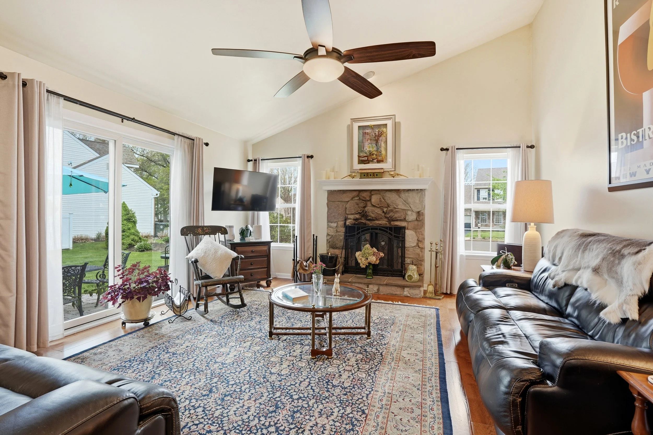 Living room with large windows, a stone fireplace, black leather sofas, a wooden rocking chair, a glass coffee table, a patterned area rug, potted plants, a TV on a stand, and decor items including framed art and candles.