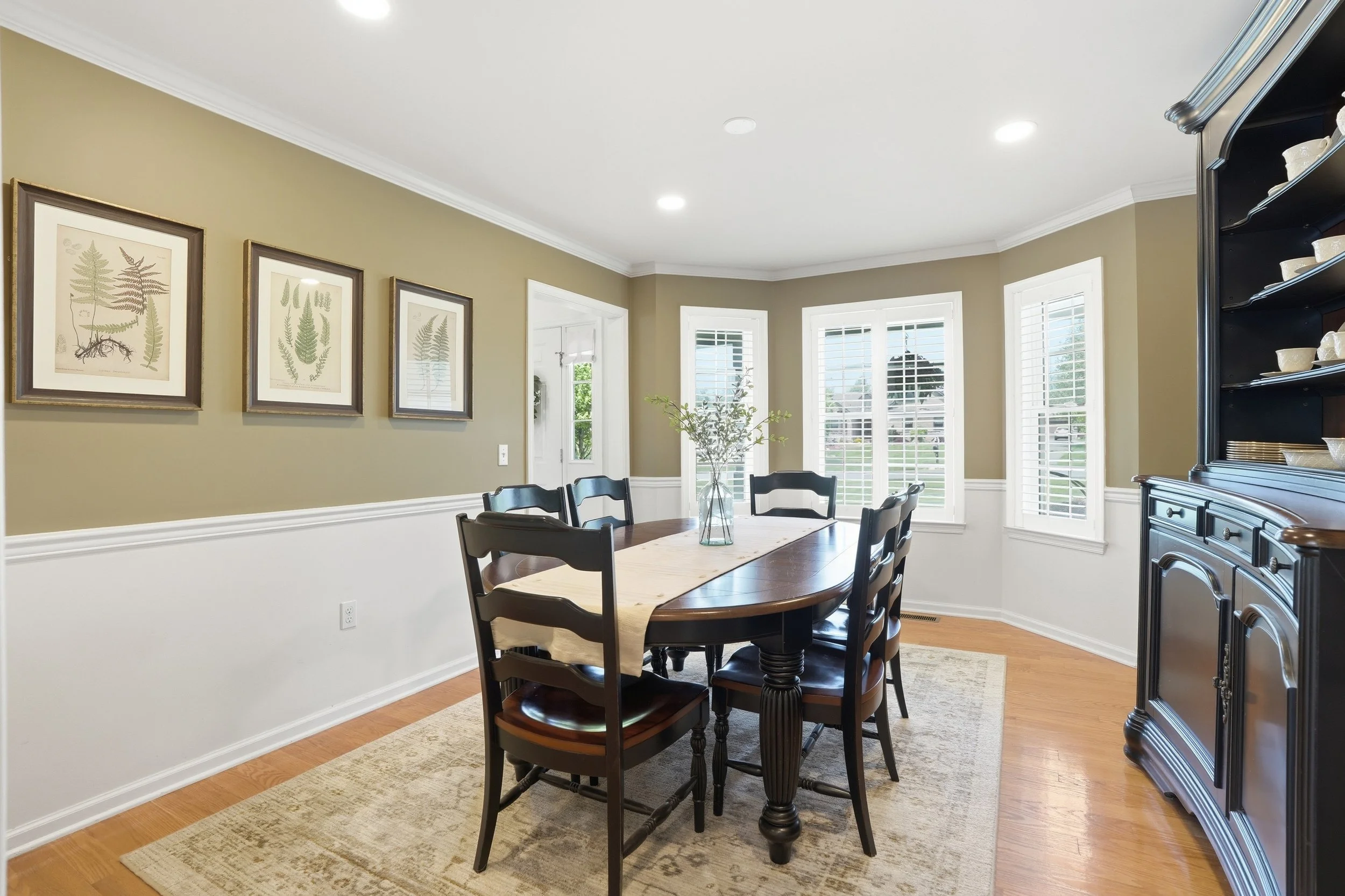 A dining room with a wooden table and six black chairs, a black china cabinet on the right, and three large windows with white blinds and plantation shutters, beige walls with white trim, framed botanical prints on the wall, and a plant in a glass va