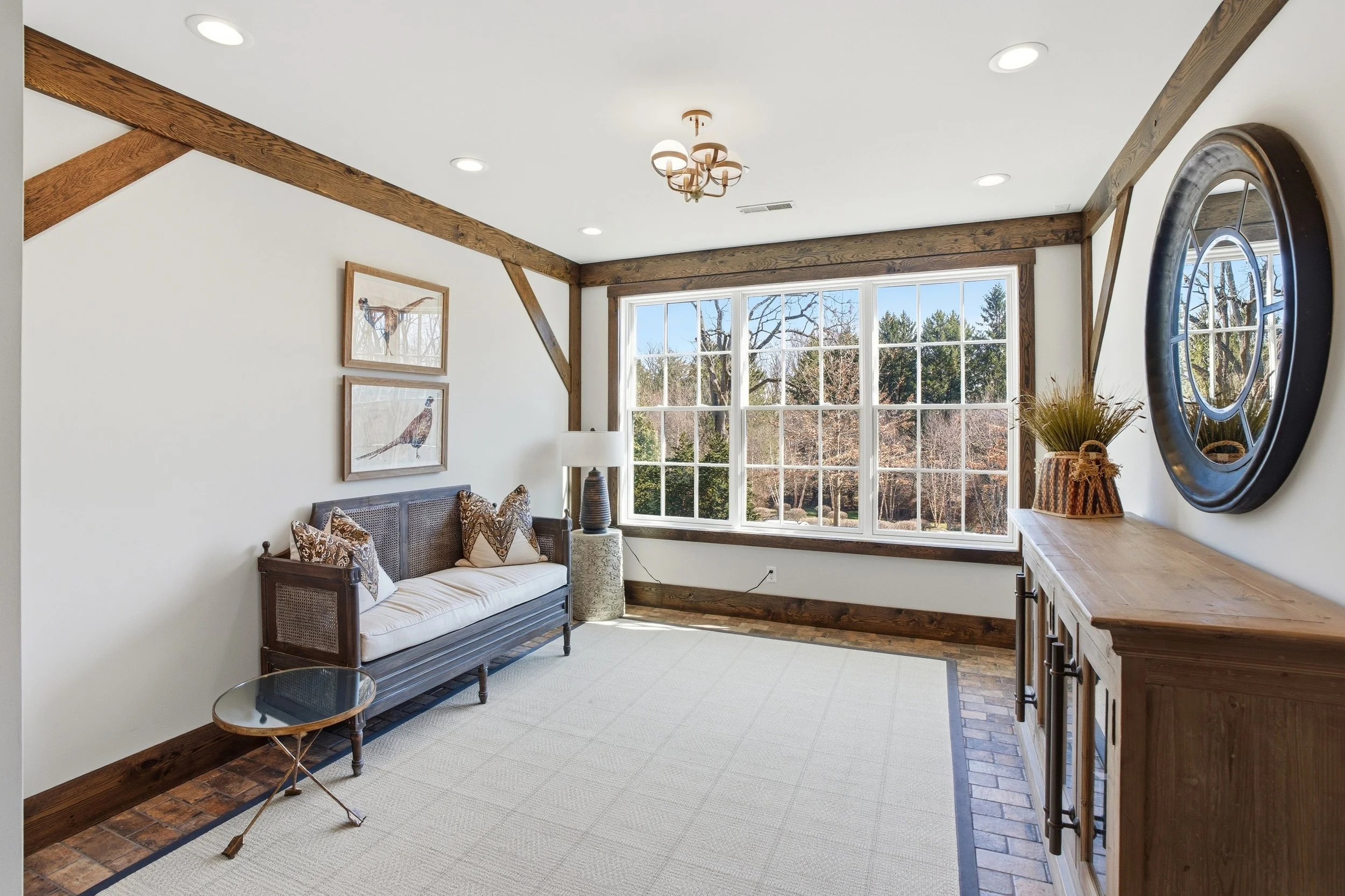 Interior of a living room with large window, wooden beams, a sofa with patterned pillows, a side table with a lamp, a round coffee table, and a wall mirror, with trees visible outside.