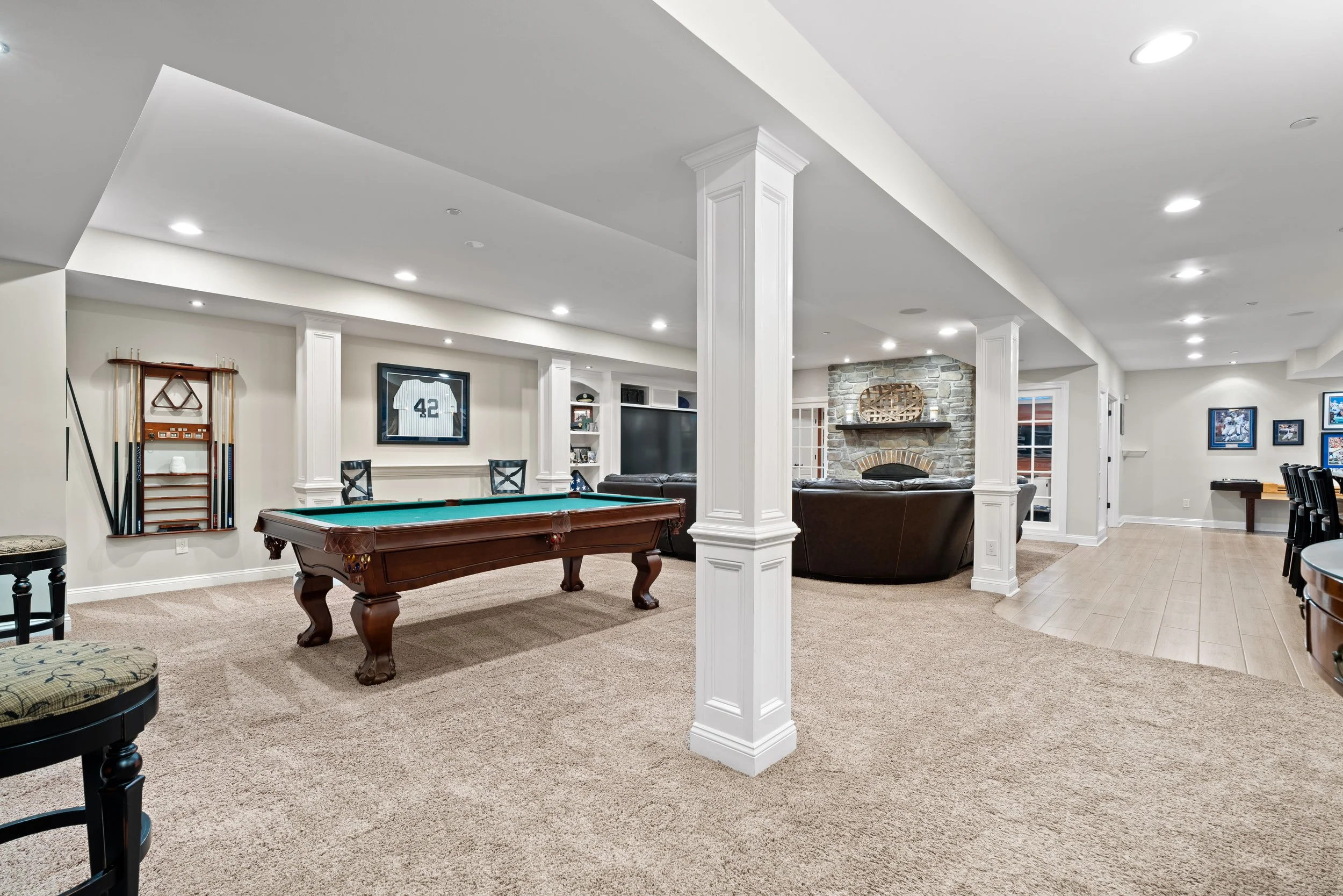 A spacious recreational room with a pool table, a stone fireplace, and framed sports jerseys and pictures on the walls, illuminated by recessed ceiling lights.