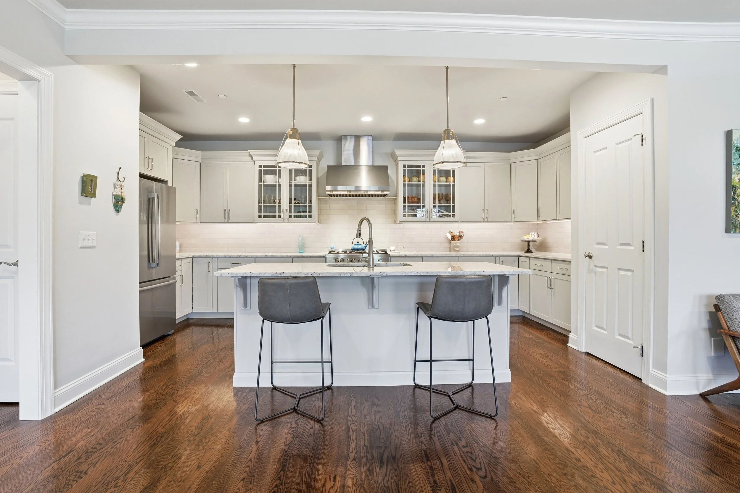 Modern white kitchen with a kitchen island, two black chairs, stainless steel refrigerator, stainless steel range hood, white cabinetry with glass doors, hardwood floor, and pendant lighting.