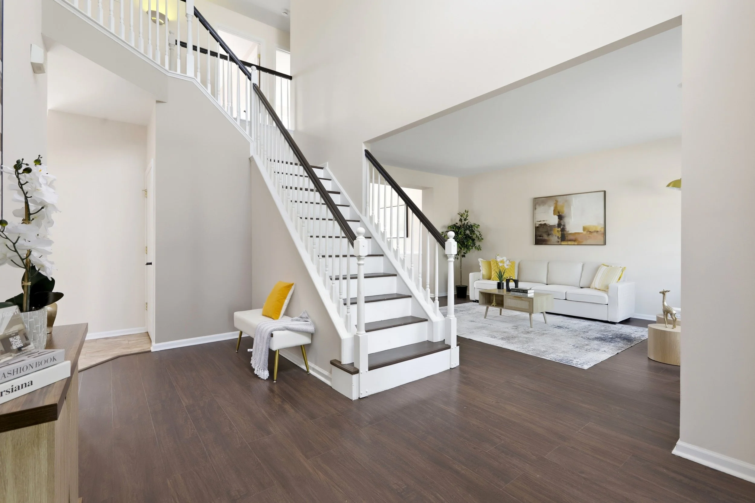 Living room with white sofa, yellow pillows, modern artwork, staircase with white and black railing, potted plant, and dark wood floors.
