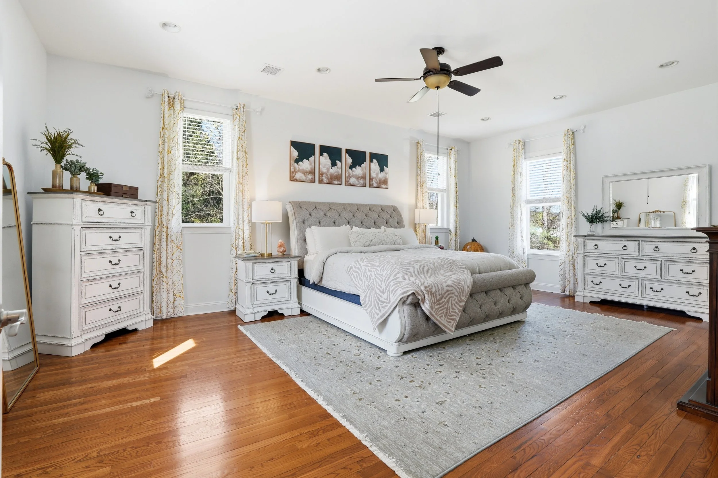 Bright bedroom with white walls, hardwood floors, a gray upholstered bed with white bedding, two white dressers, and four framed cloud pictures above the bed. Windows with white blinds and yellow-patterned curtains, two white bedside lamps, a corner 
