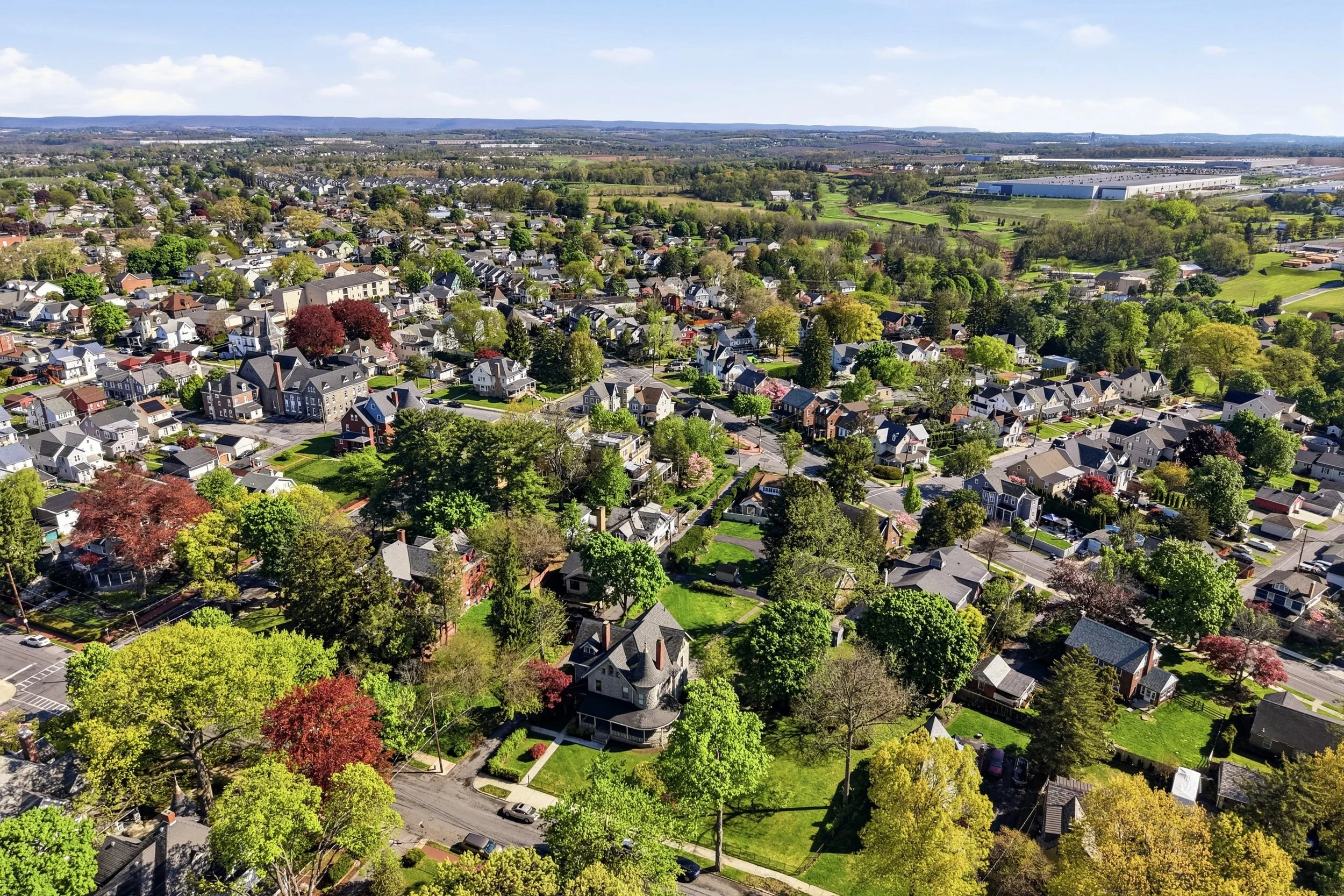 An aerial view of a suburban neighborhood with numerous houses, trees, and green spaces under a clear blue sky.