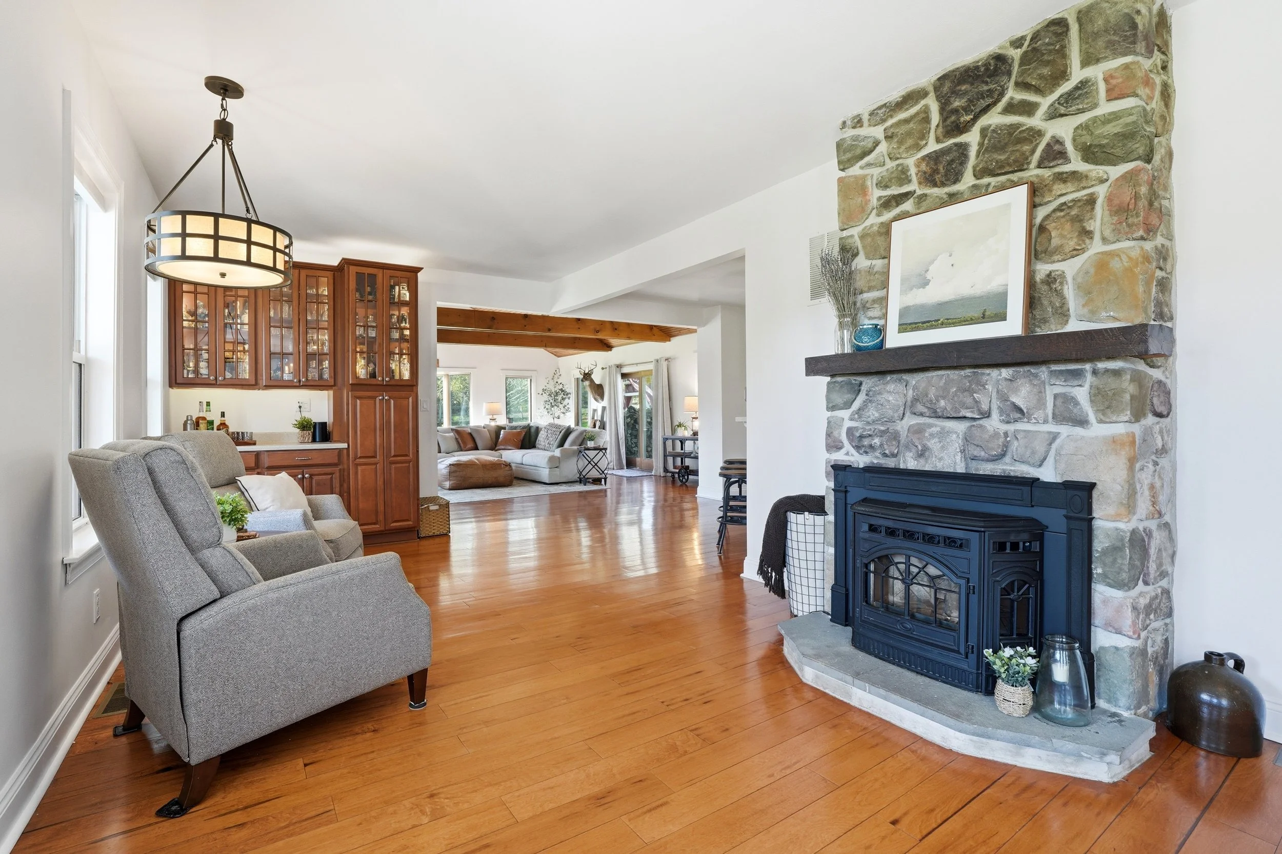 A living room with beige armchairs, a stone fireplace with a framed picture and decorative vases on the mantel, wooden floors, and a background area with a beige sofa, deer head mounted on the wall, and large windows.