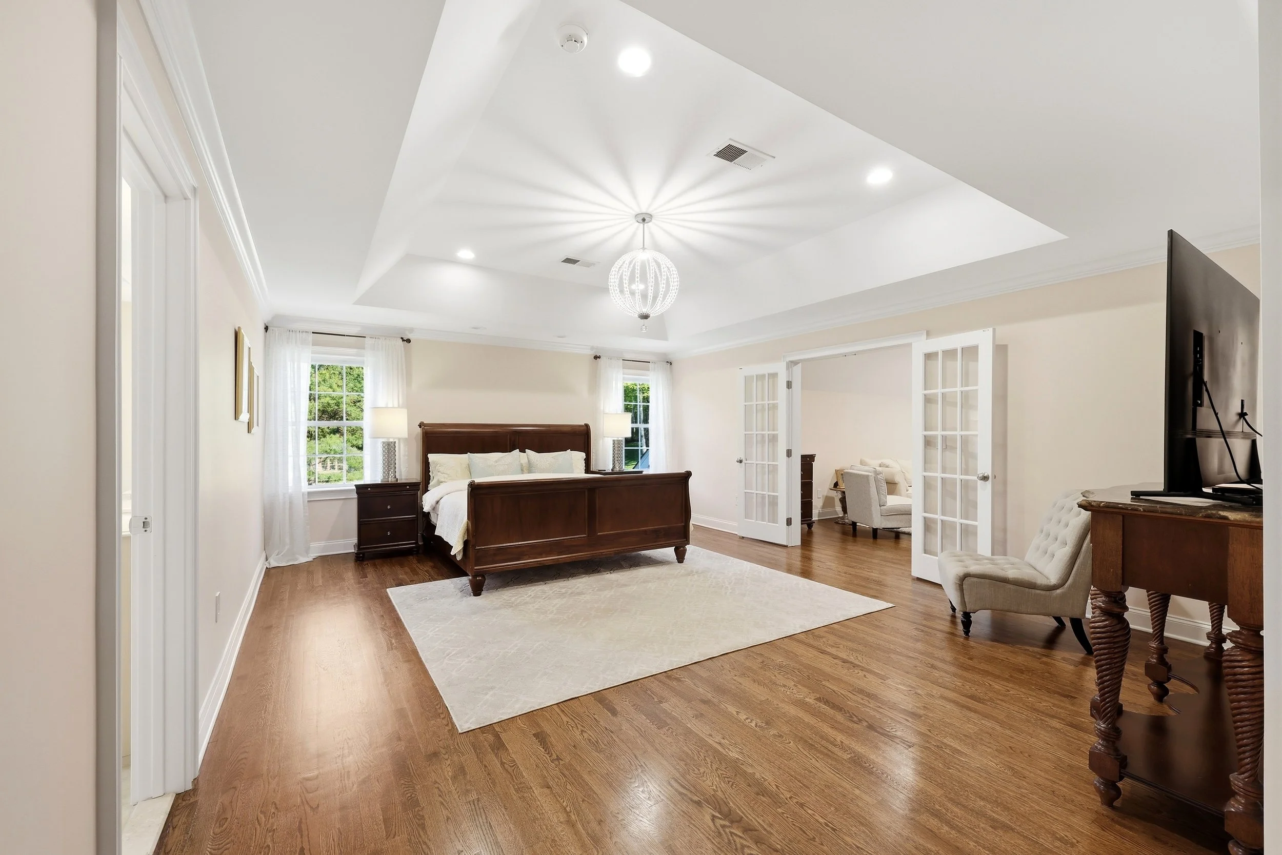 Living room with a wooden bed frame, white bedding, two nightstands, lamps, and hardwood flooring. French doors lead to another room, and a television is visible on the right. Large windows with curtains provide natural light, and a modern chandelier