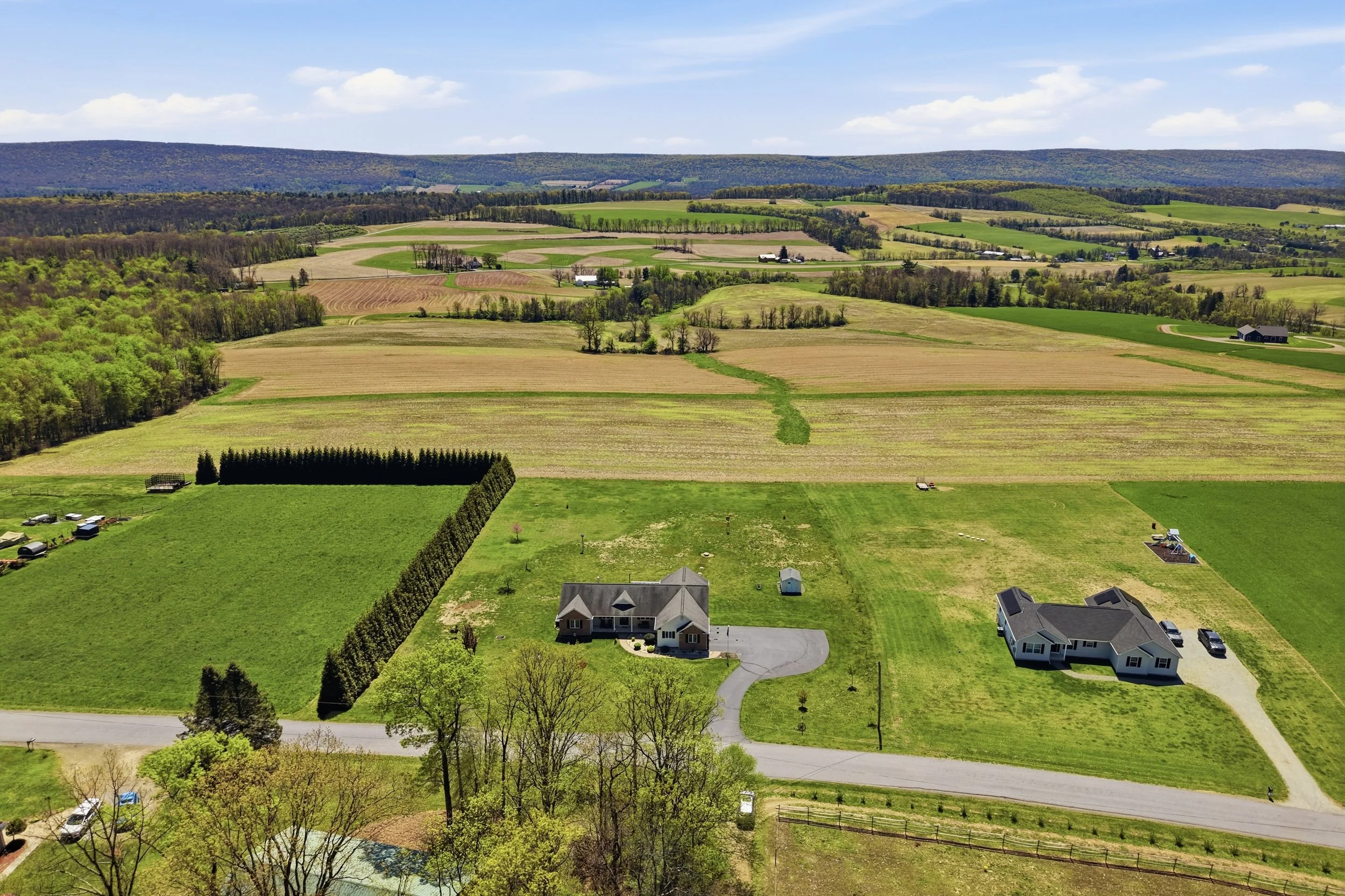 Aerial view of rural farmland with houses, trees, green fields, and hills in the distance under a blue sky with clouds.