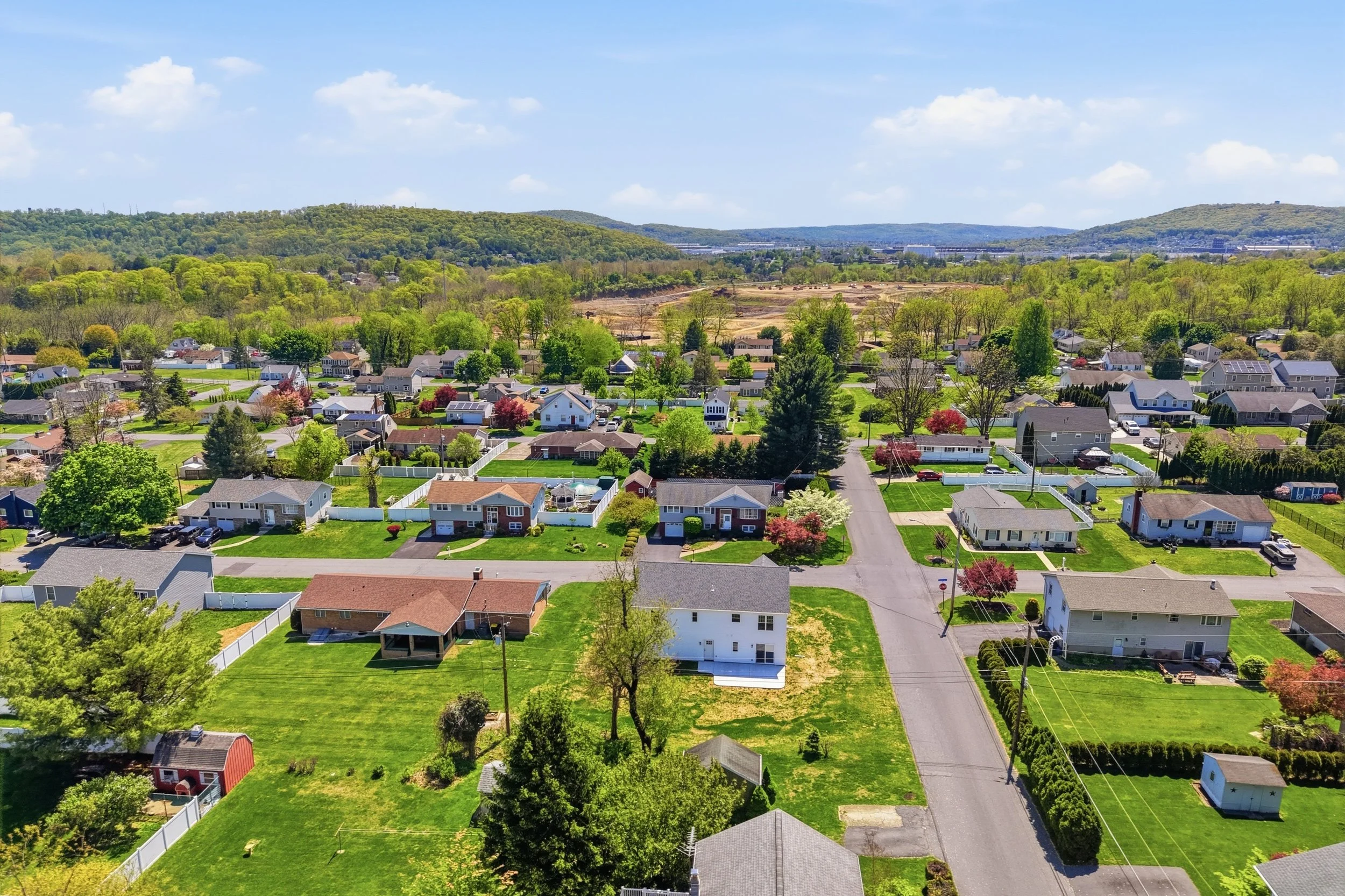 Aerial view of a suburban neighborhood with houses, lawns, and trees, with green hills and a blue sky in the background.