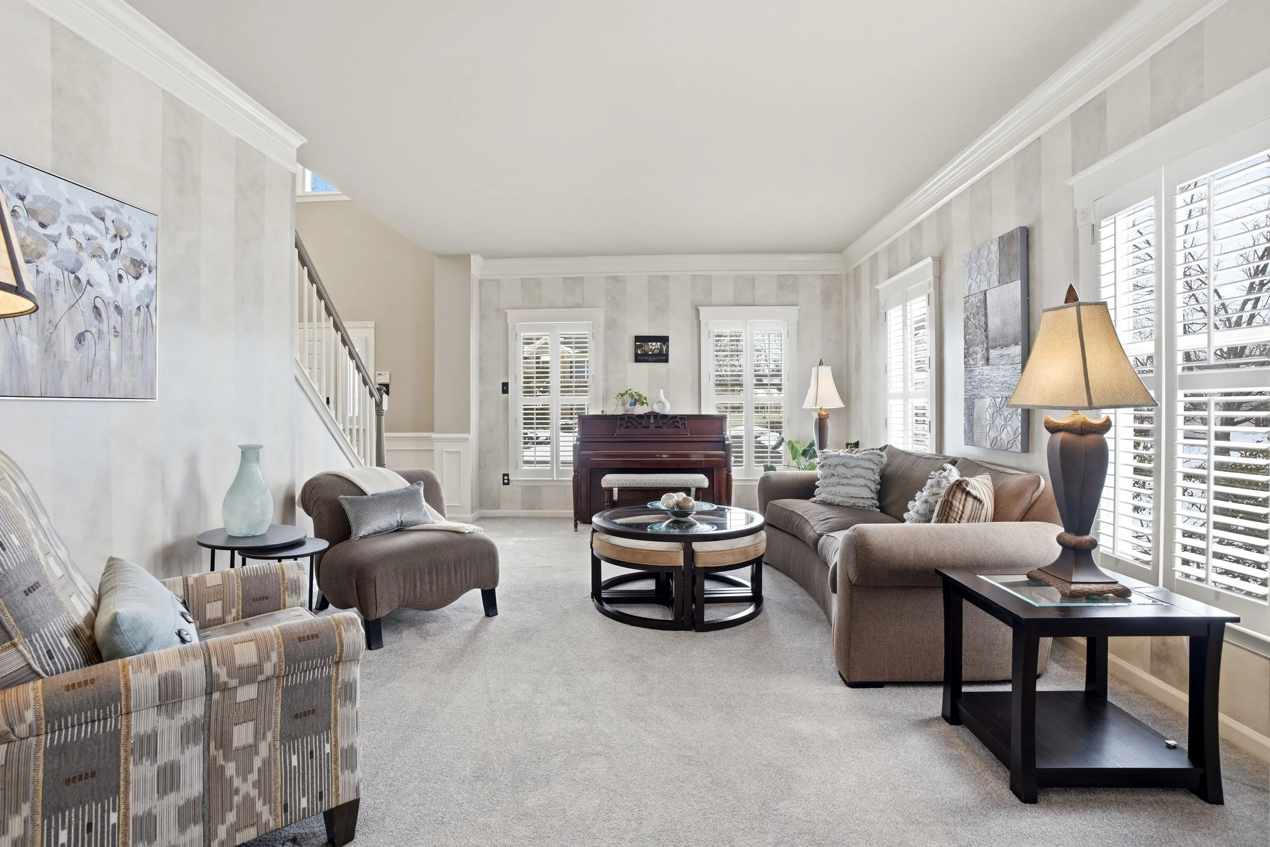 Living room with beige sofas, black side tables, a wooden piano, white trimmed windows with blinds, wall art, and lamps.