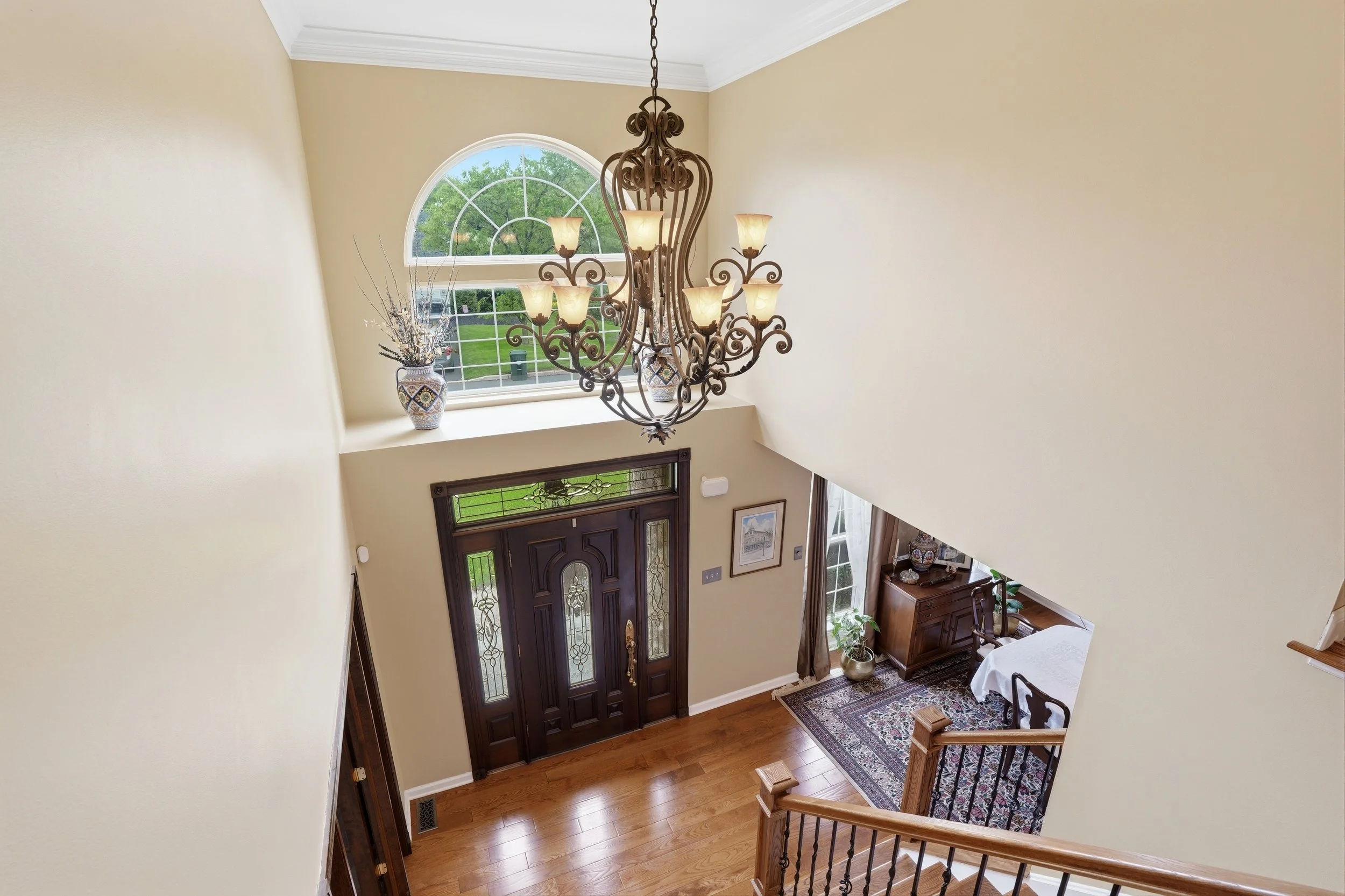 Interior view of a house entrance with a dark wooden front door, a large arched window above, a chandelier hanging from the ceiling, a patterned rug on the wooden floor, and a staircase with wooden handrails. There are decorative vases with dried bra