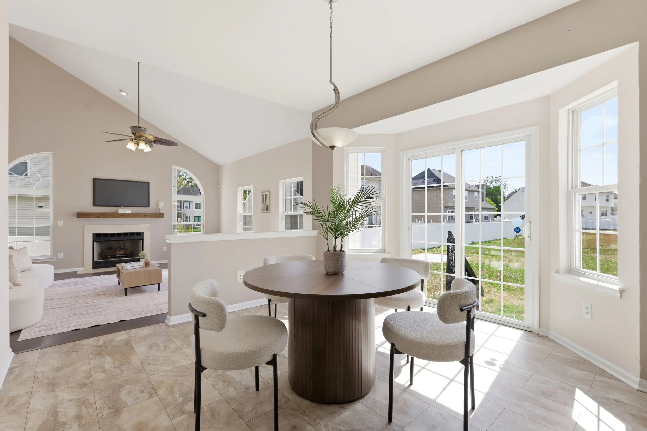 Bright living room with large windows, a white sofa, a wooden coffee table, a wall-mounted TV above a fireplace, ceiling light, and a ceiling fan, with a view of neighboring houses outside.