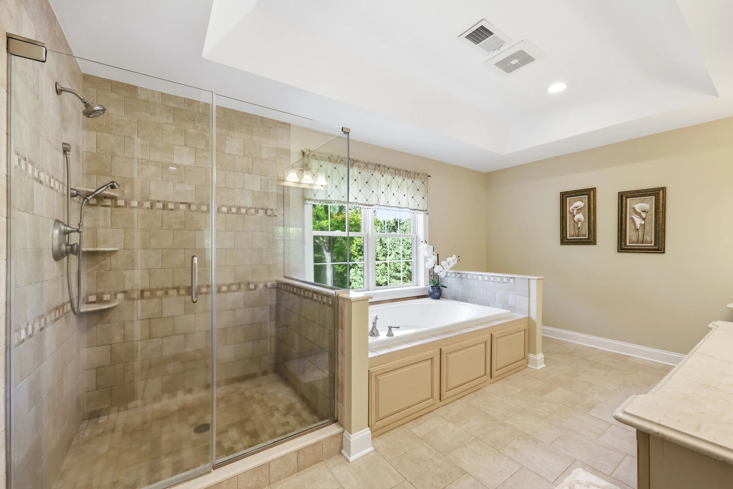 Bathroom with glass-enclosed shower, beige tiled walls, a bathtub next to a window with curtains, and decorative framed art on the wall.