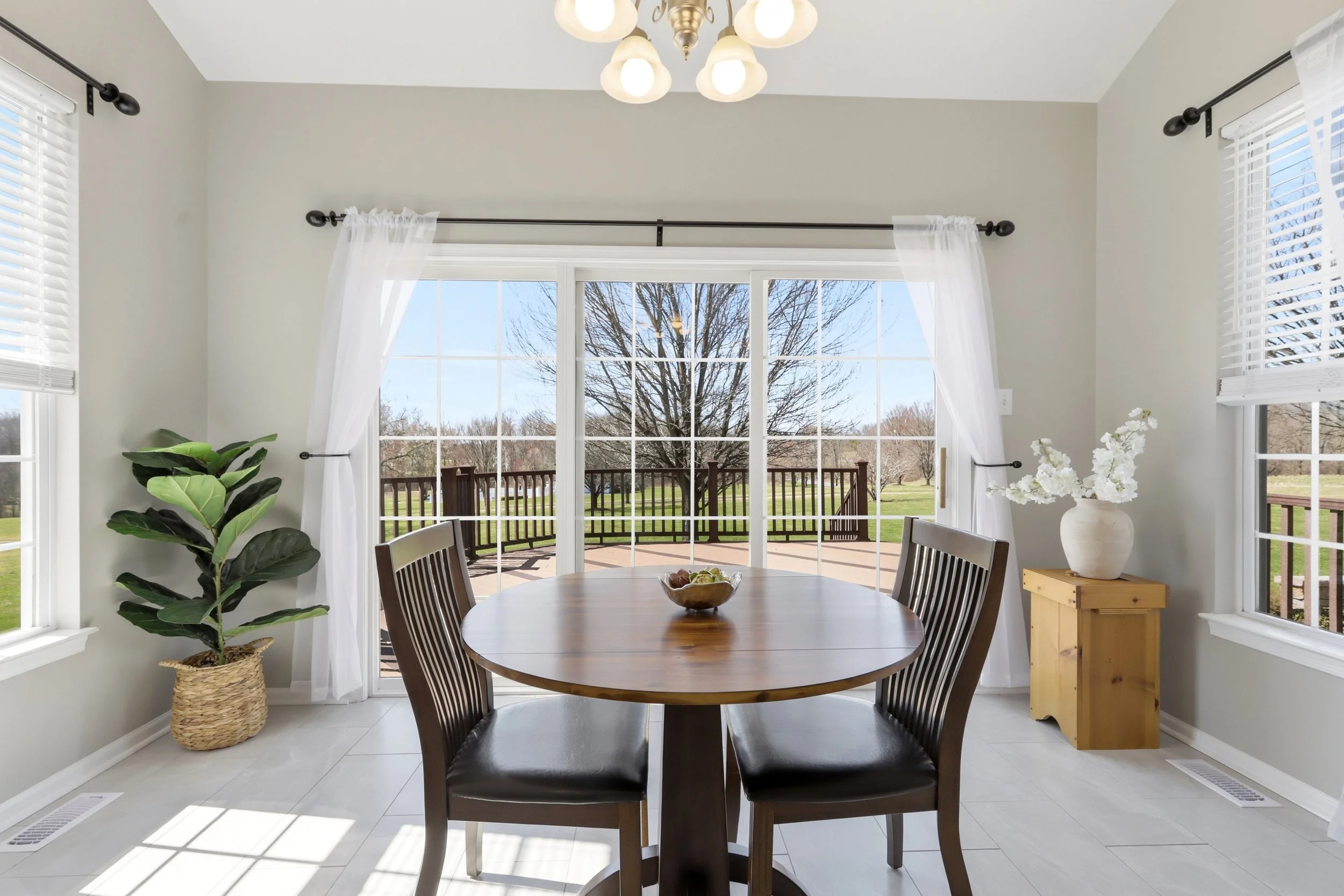 Sunlit dining area with a round wooden table and four black chairs, large windows with white blinds, white curtains, a potted plant, a white vase with flowers, and a sliding glass door leading to a deck with trees outside.