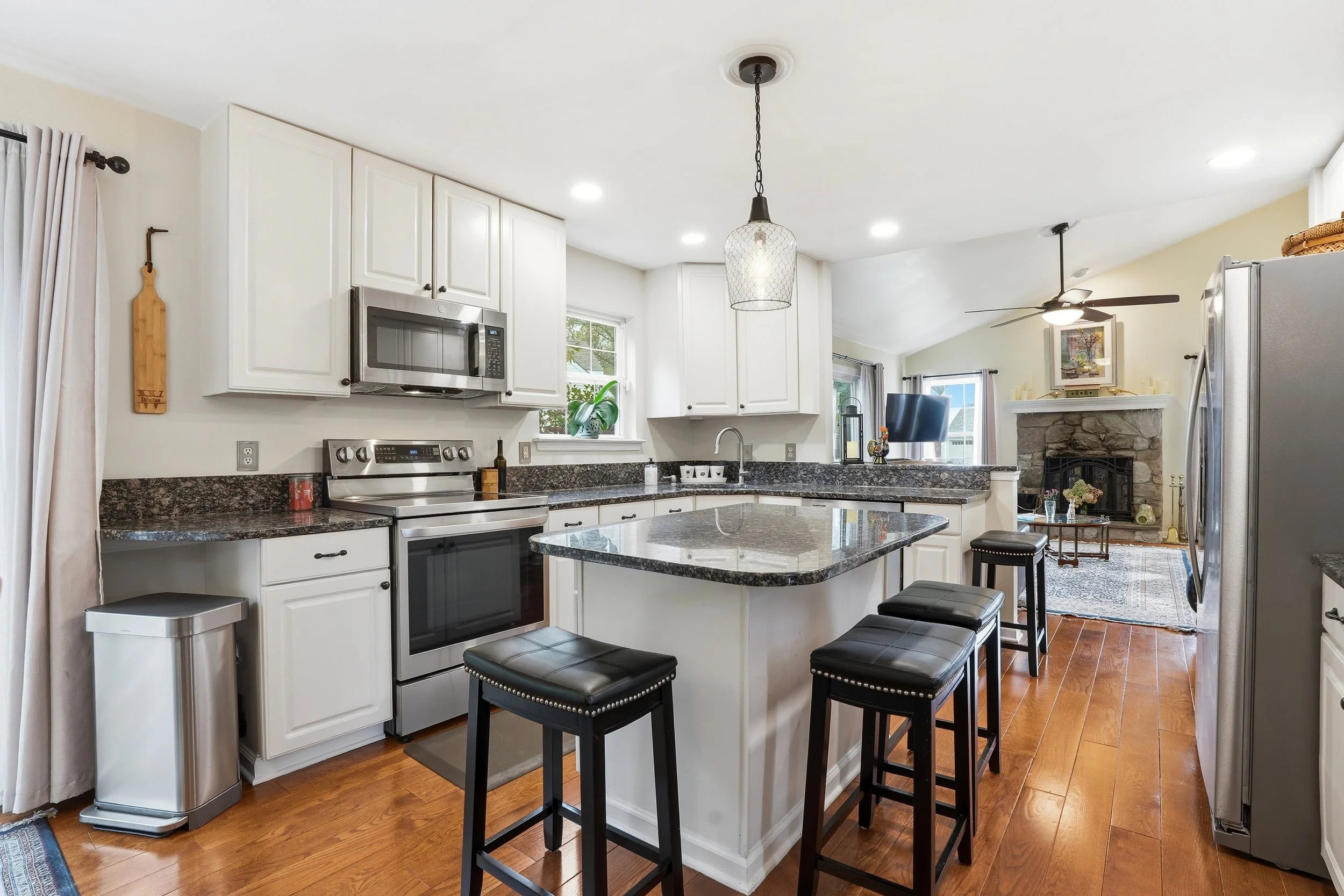 A modern kitchen with white cabinets, black granite countertops, a stainless steel oven, microwave, and a refrigerator. There are three black barstools at a breakfast bar and a view of a living area with a stone fireplace and a ceiling fan.