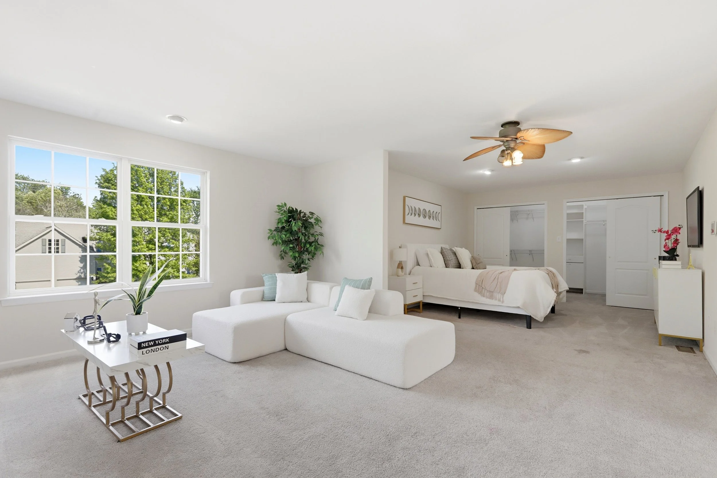 Bright living room with large window, white sofa, green plant, wall art, ceiling fan, and open closet with sliding doors.