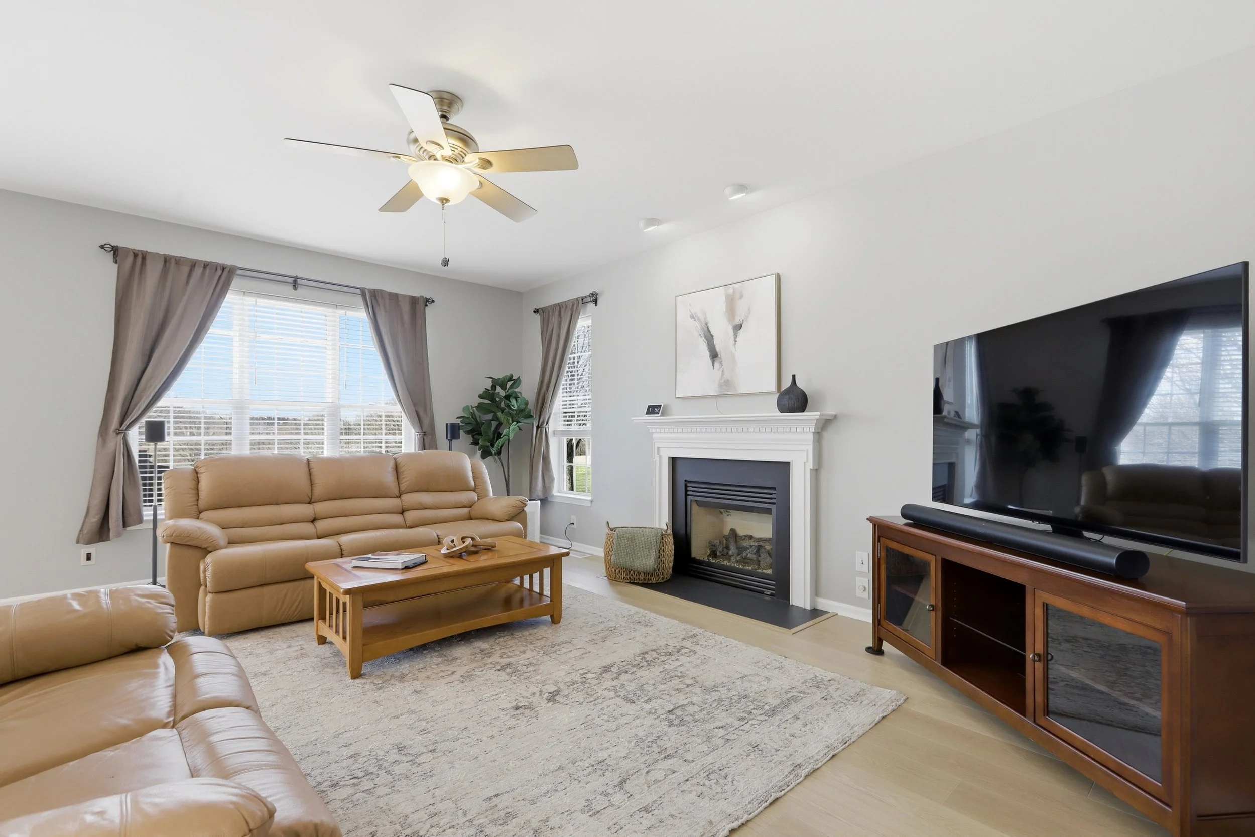 Living room with beige leather sofa, wooden coffee table, fireplace, large window with beige curtains, TV on a wooden stand, ceiling fan with light, artwork above fireplace, houseplant near window, and beige carpet.