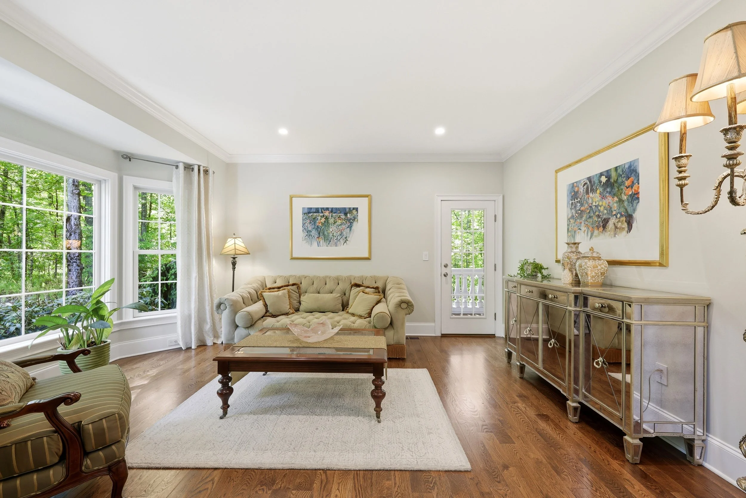 A living room with large windows showing greenery outside, a beige tufted sofa with pillows, a wooden coffee table, a mirrored sideboard with vases, paintings on the walls, a floor lamp, and hardwood floors.