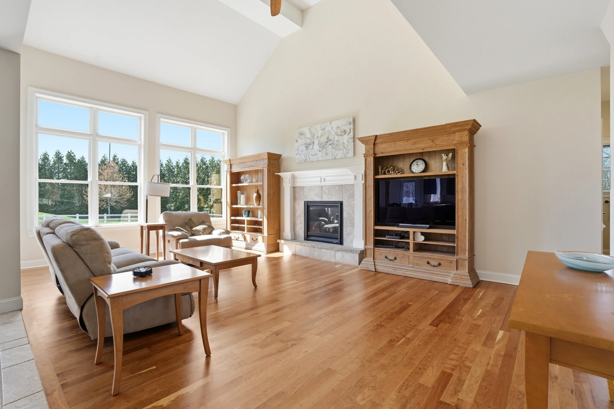 Living room with large windows, beige sofa, wooden coffee table, and wooden entertainment center with a flat-screen TV. There is a fireplace with a white mantel and decorative items. The room has hardwood floors and cream-colored walls, with sunlight