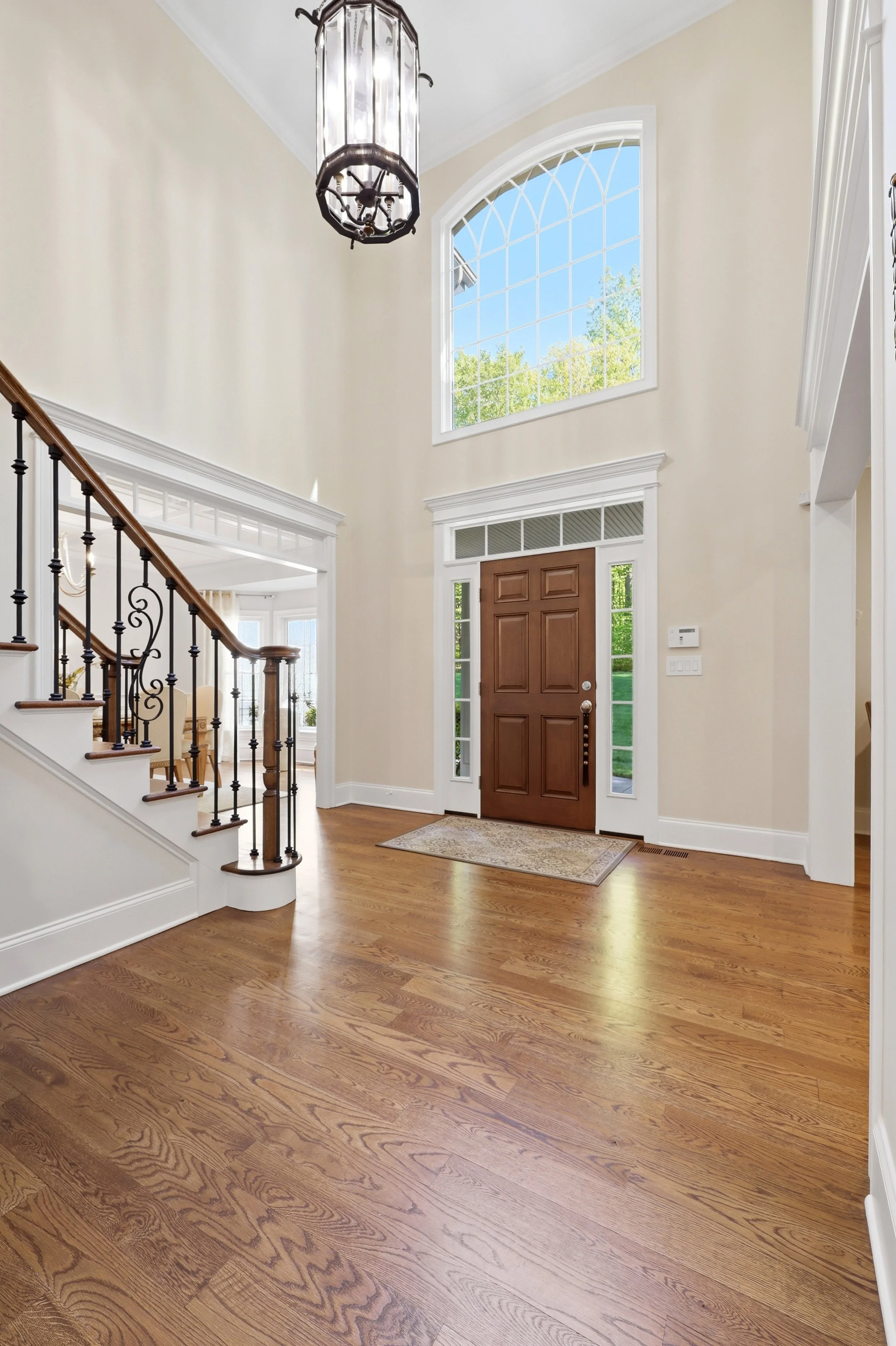 Entryway with wooden front door, large window, staircase with black wrought iron railing, and chandelier, hardwood floors, and beige walls.