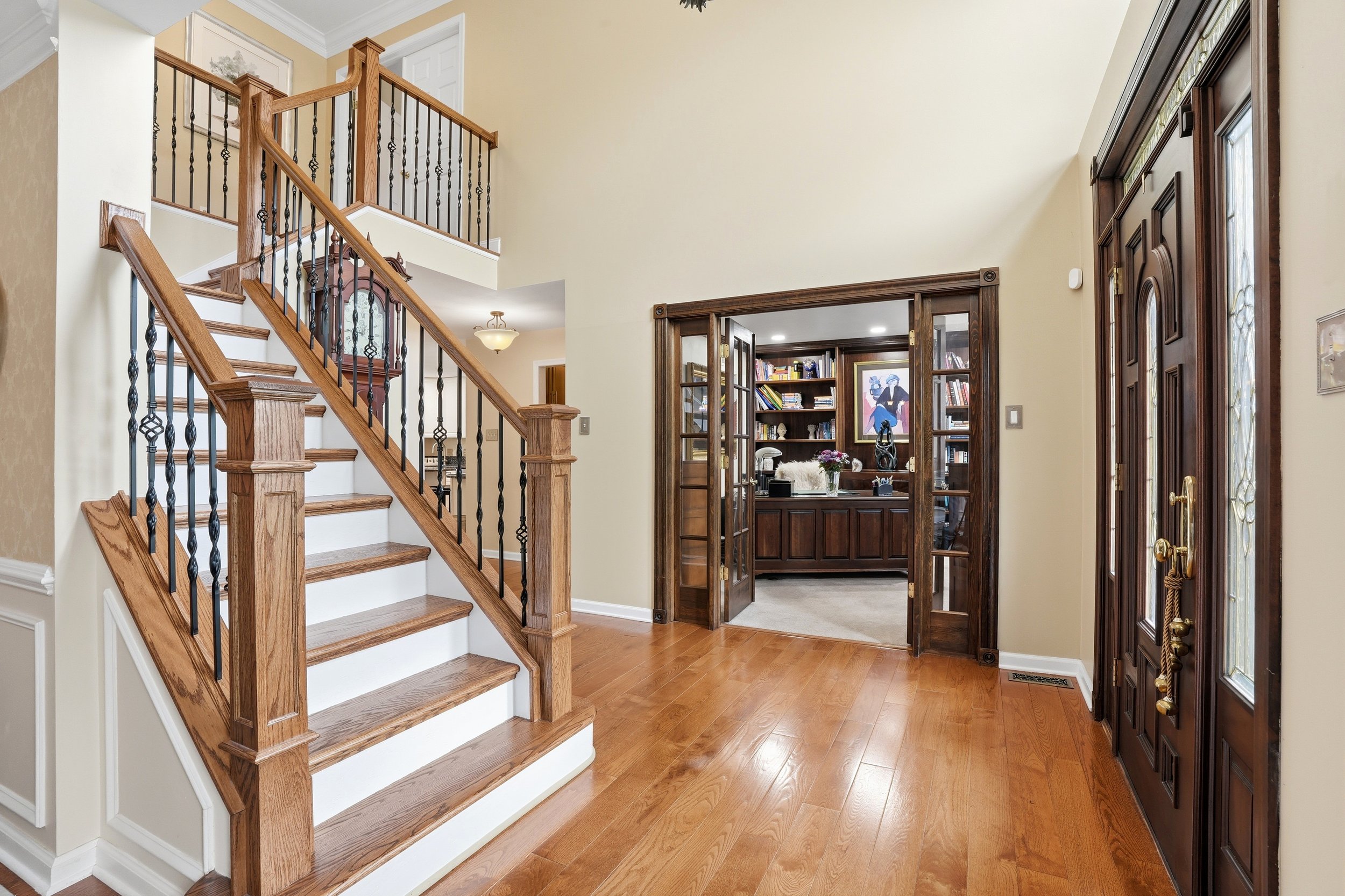 Interior view of a home with a wooden staircase on the left, wood flooring, and an open doorway on the right leading to a room with bookshelves and a decorative painting. The wall color is light cream, and there are dark wood trim and doors.