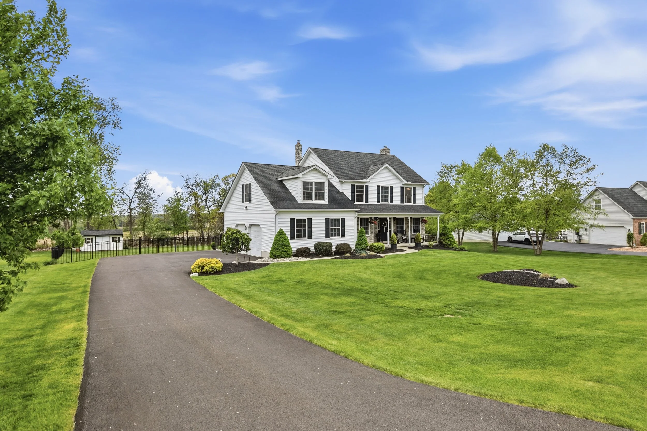 A large white house with black shutters, a gray roof, and a porch, surrounded by green trees and well-maintained lawns, with a curved asphalt driveway leading up to the house under a partly cloudy blue sky.