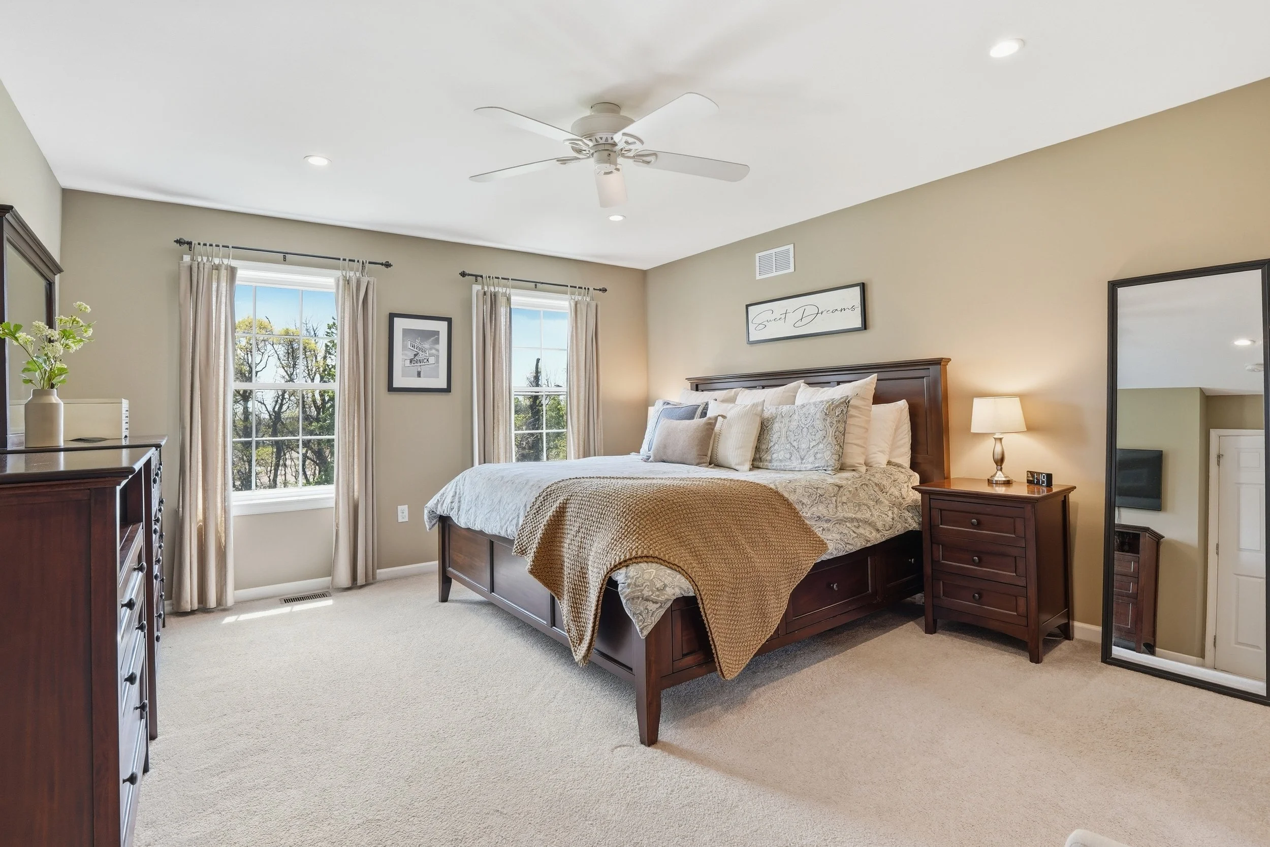 A neatly made bed with beige and white bedding in a spacious bedroom with beige walls, two windows with cream curtains, a mirror, a bedside lamp, and framed wall art above the bed.