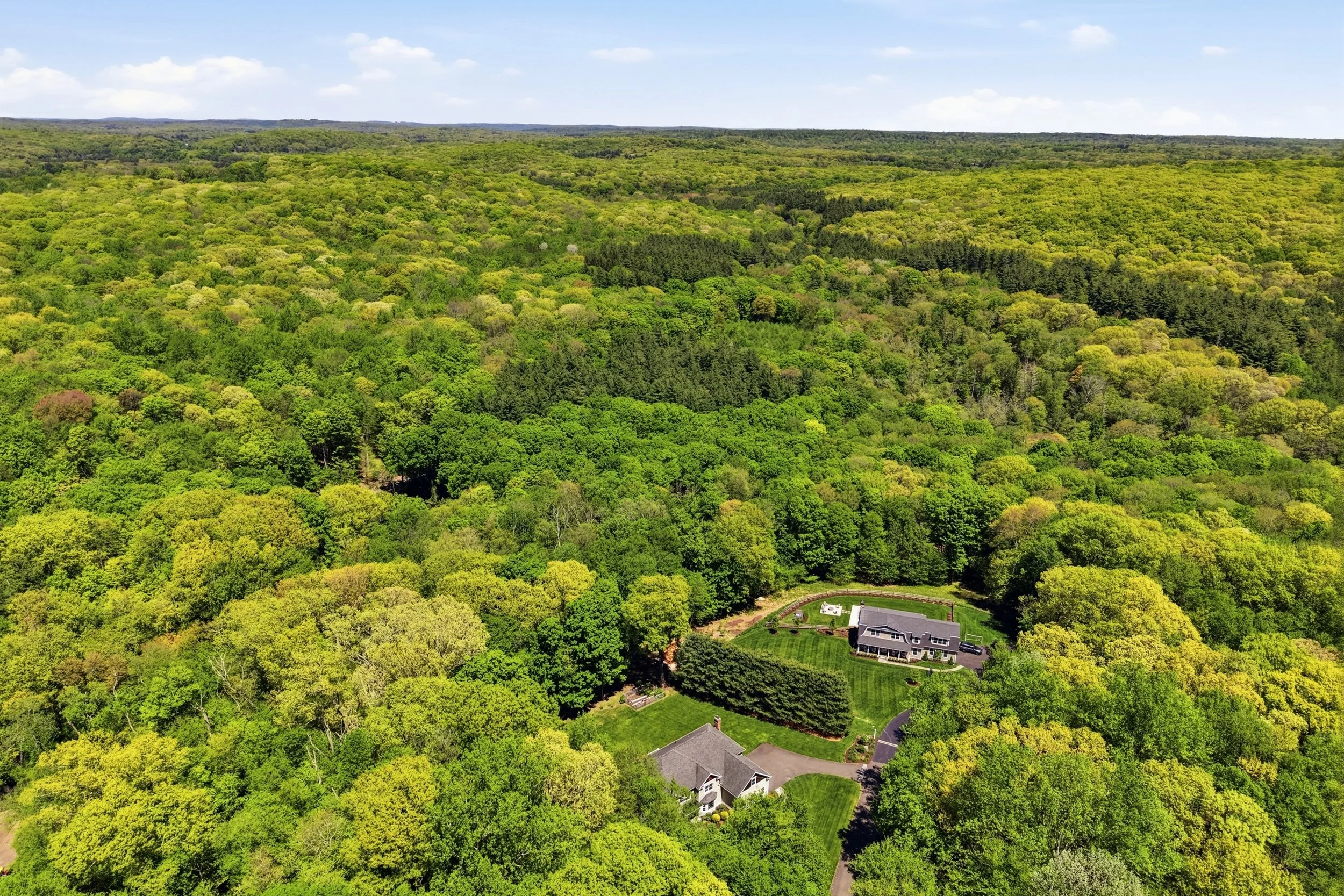 Aerial view of a large house surrounded by lush green trees in a forested area under a blue sky with scattered clouds.