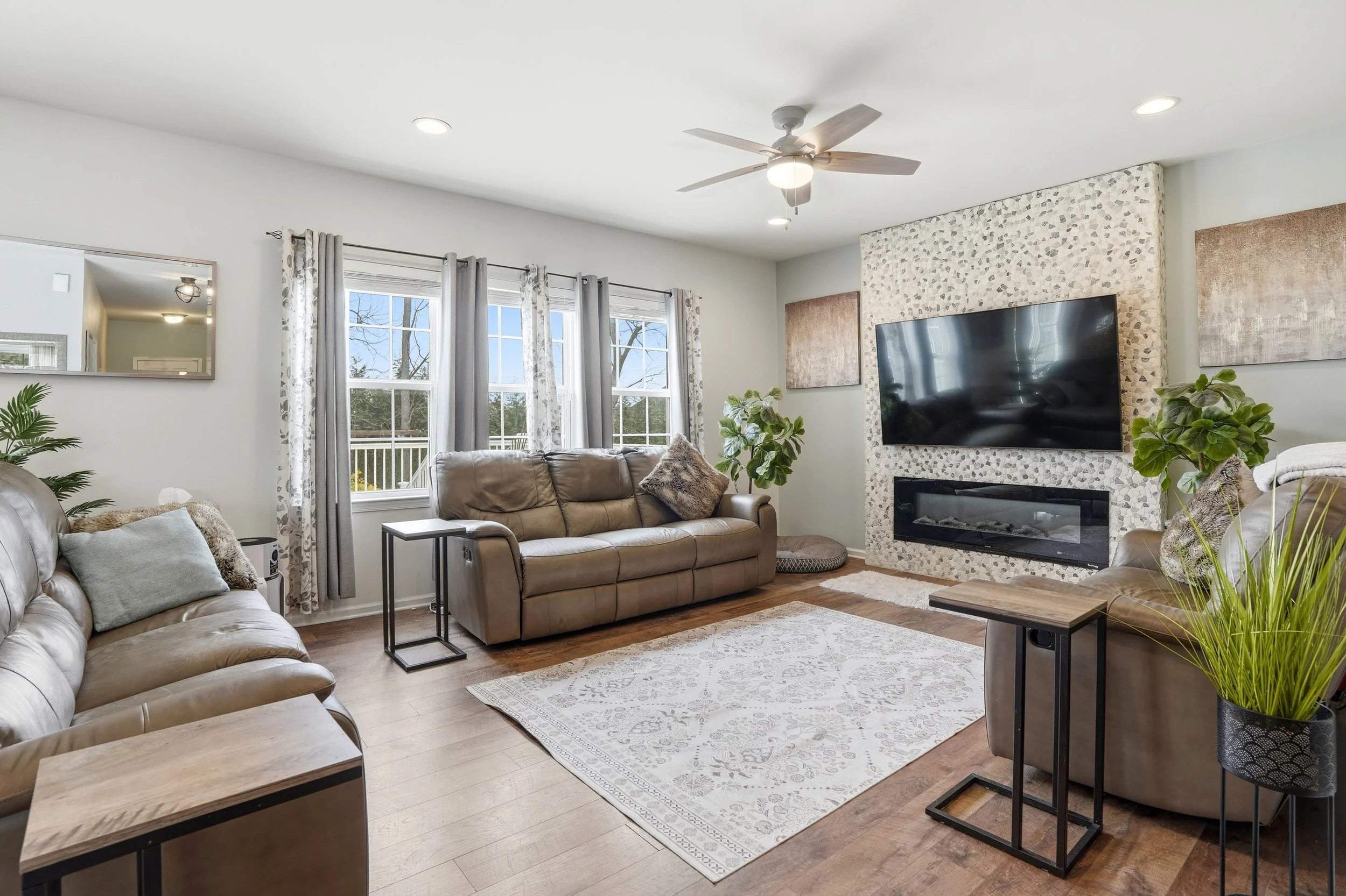 A living room with three large windows with curtains, a leather sofa, a leather recliner, a fireplace with a large flat-screen TV mounted above, various plants, and abstract artwork on the walls.