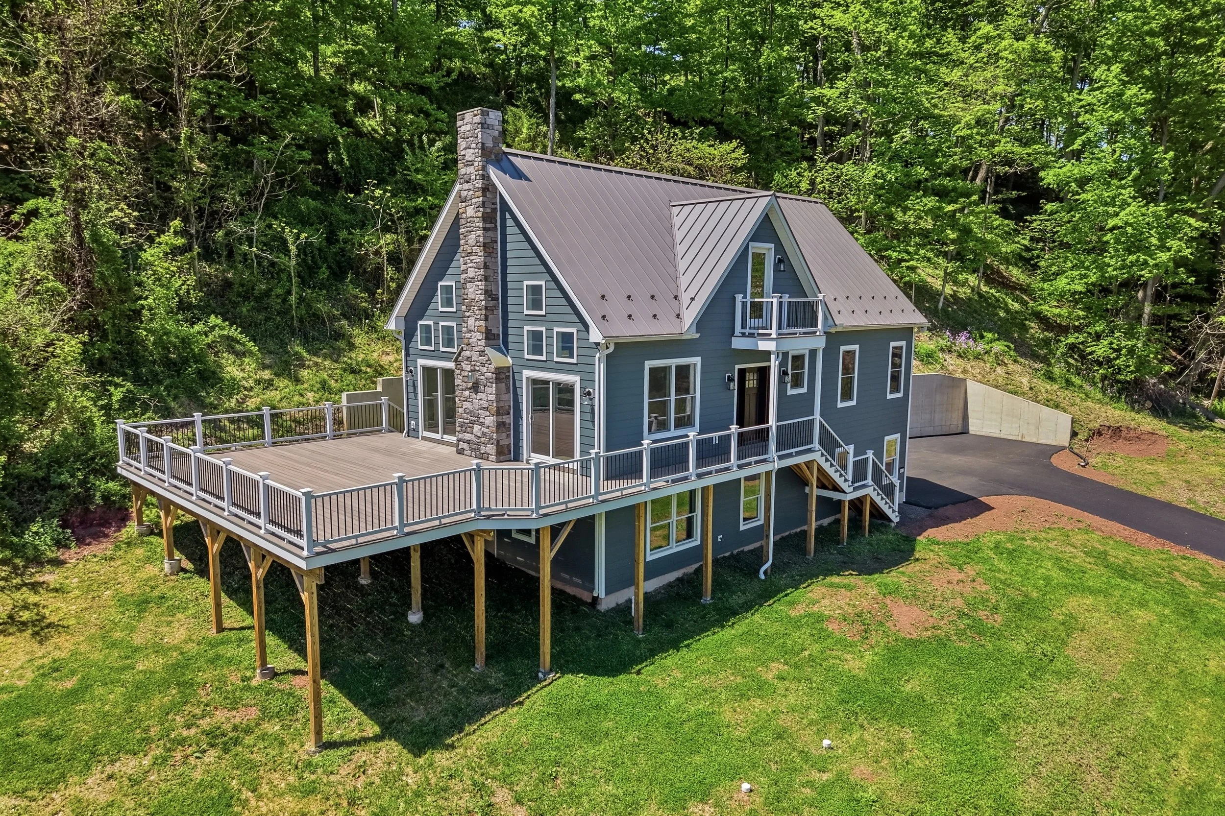 A blue house with a gray metal roof and a prominent stone chimney, featuring a large wooden deck with white railing, situated in a lush, green wooded area.