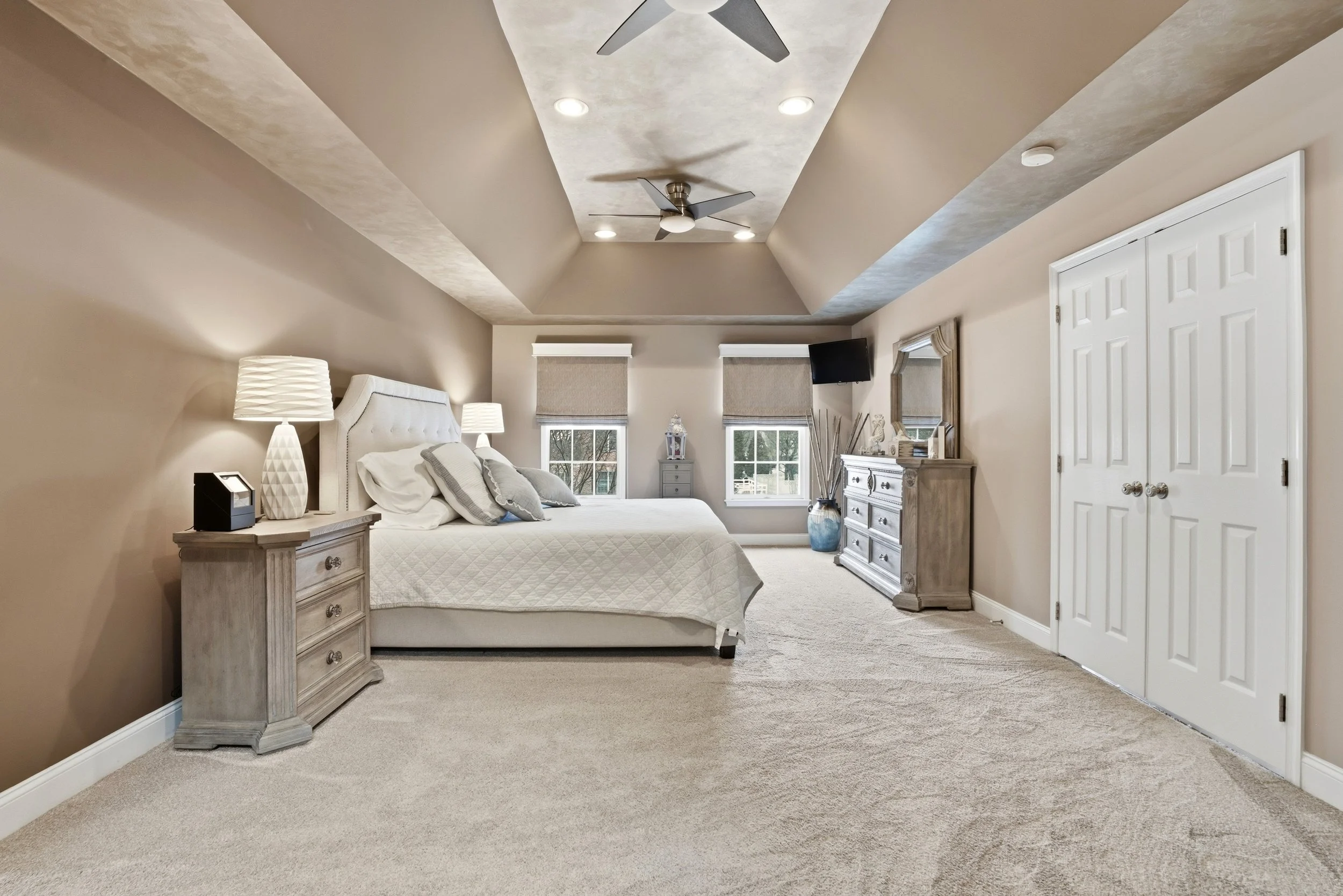 A bedroom with a light-colored bed, matching nightstands with lamps, and a dresser with a mirror. There are two small windows on the far wall, and a TV mounted near the ceiling. The room has beige walls, carpeted flooring, and a tray ceiling with two