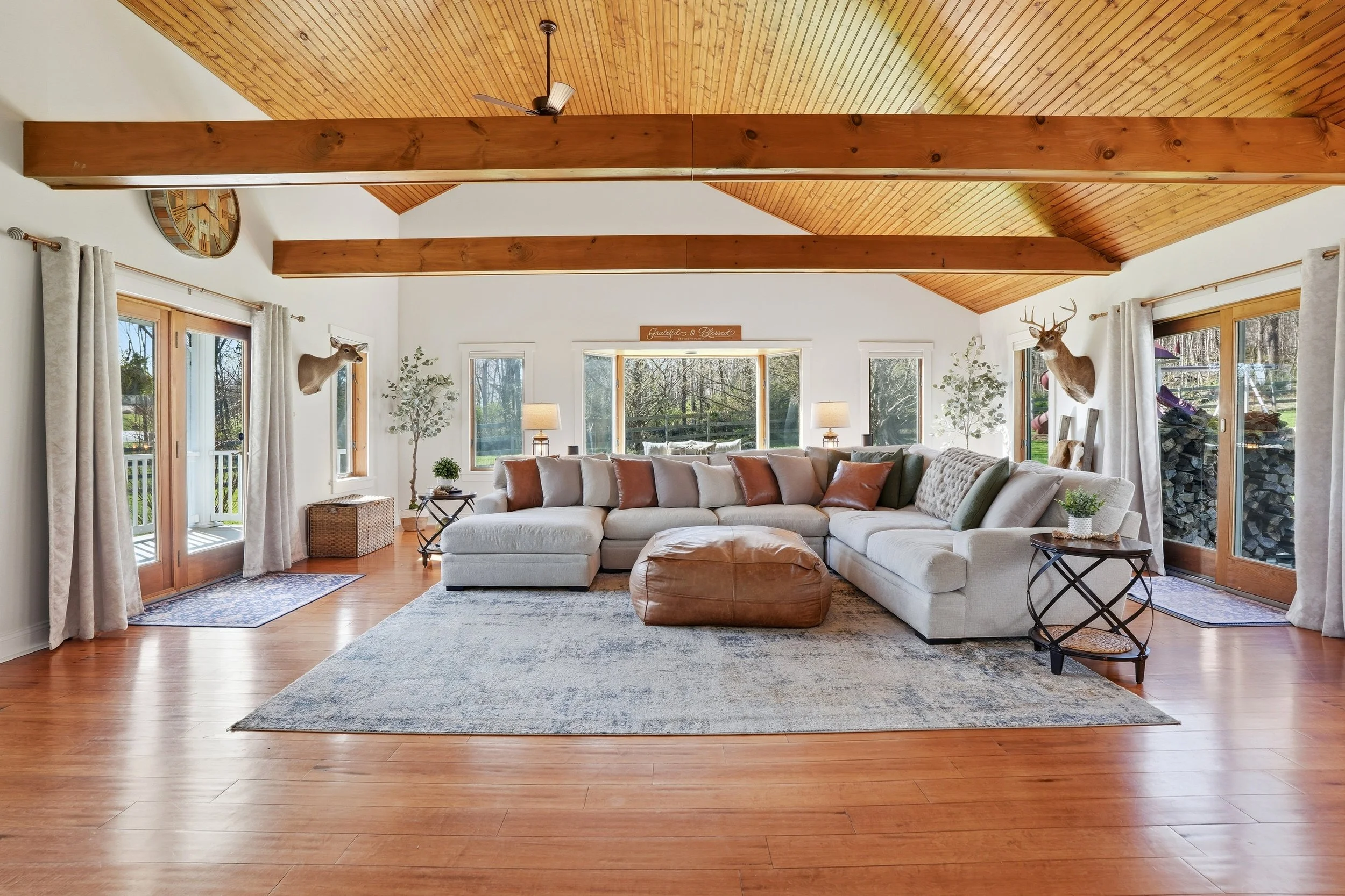 Bright living room with white sectional sofa, brown leather ottoman, and side tables with lamps. Large windows and sliding glass doors with curtains, wood-paneled vaulted ceiling with beams and ceiling fan, deer head mounted on wall, and a view of tr