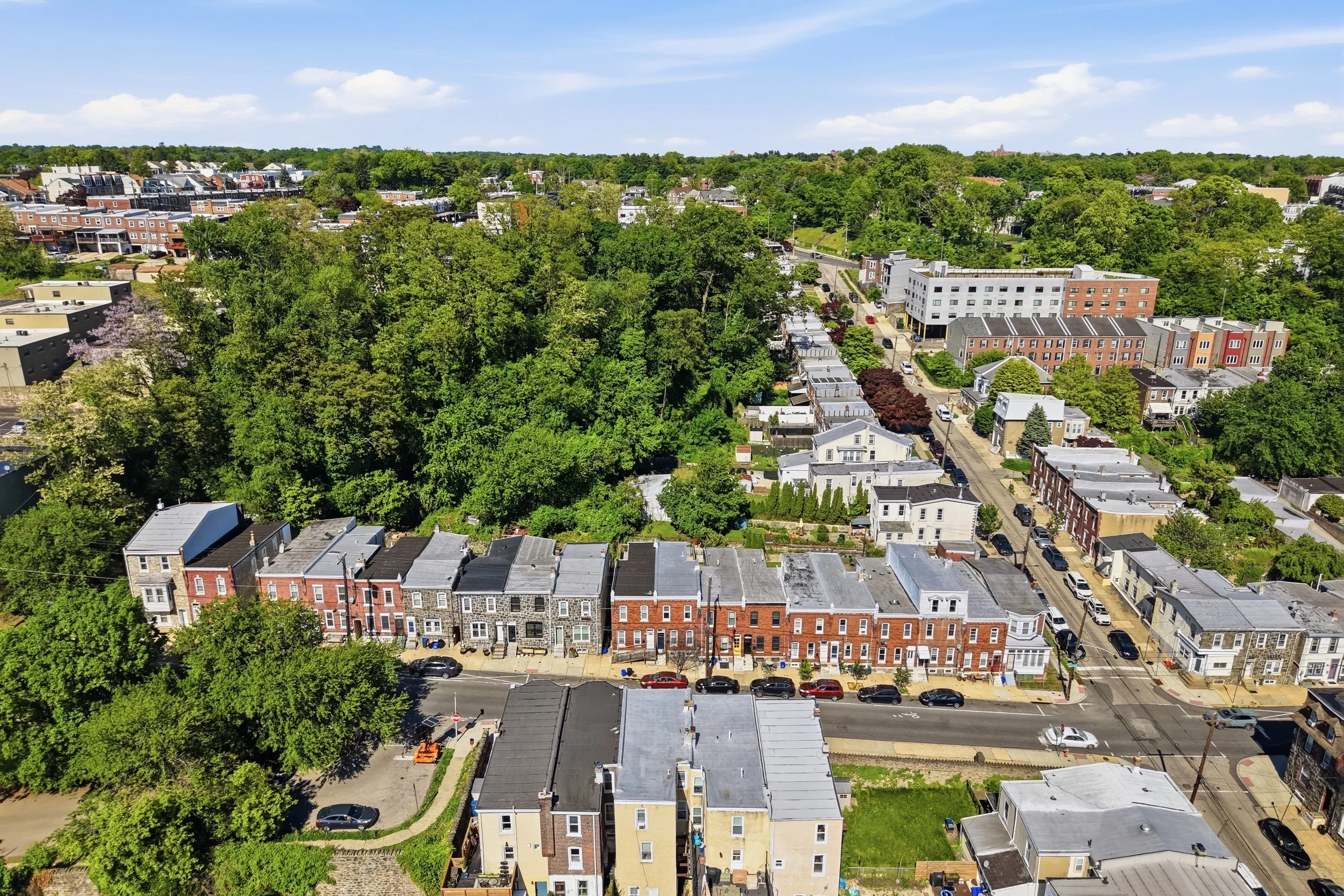 Aerial view of a city neighborhood with a mix of residential buildings and lush green trees under a blue sky with scattered clouds.