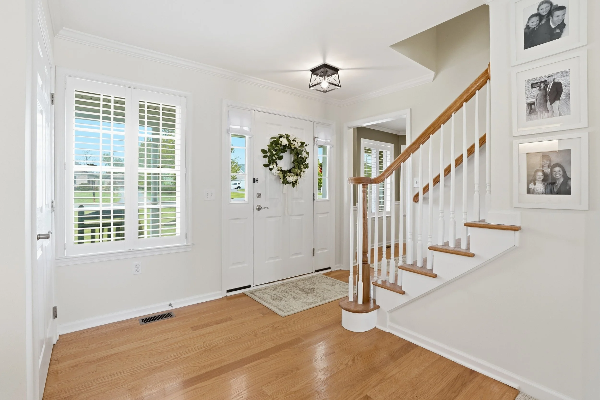 Interior of a home entryway with white walls, a front door decorated with a green wreath, a doormat, a staircase with white risers and wooden steps, framed family photos on the wall, and large windows with shutters.