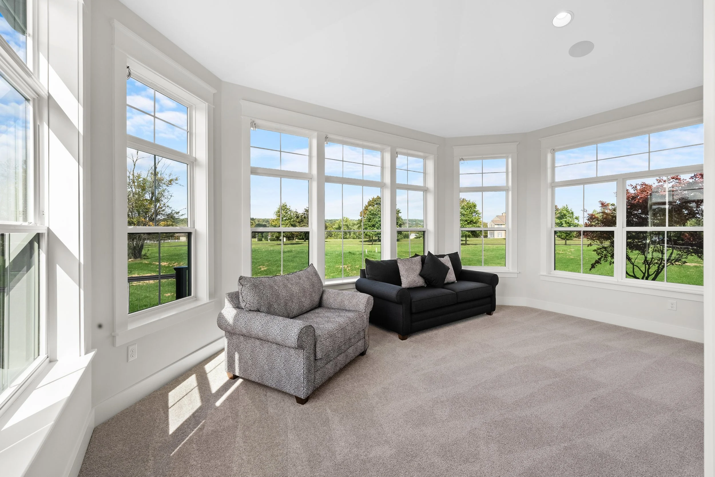 Bright living room with large windows, two couches (one black, one patterned), and a view of a green yard and trees outside.
