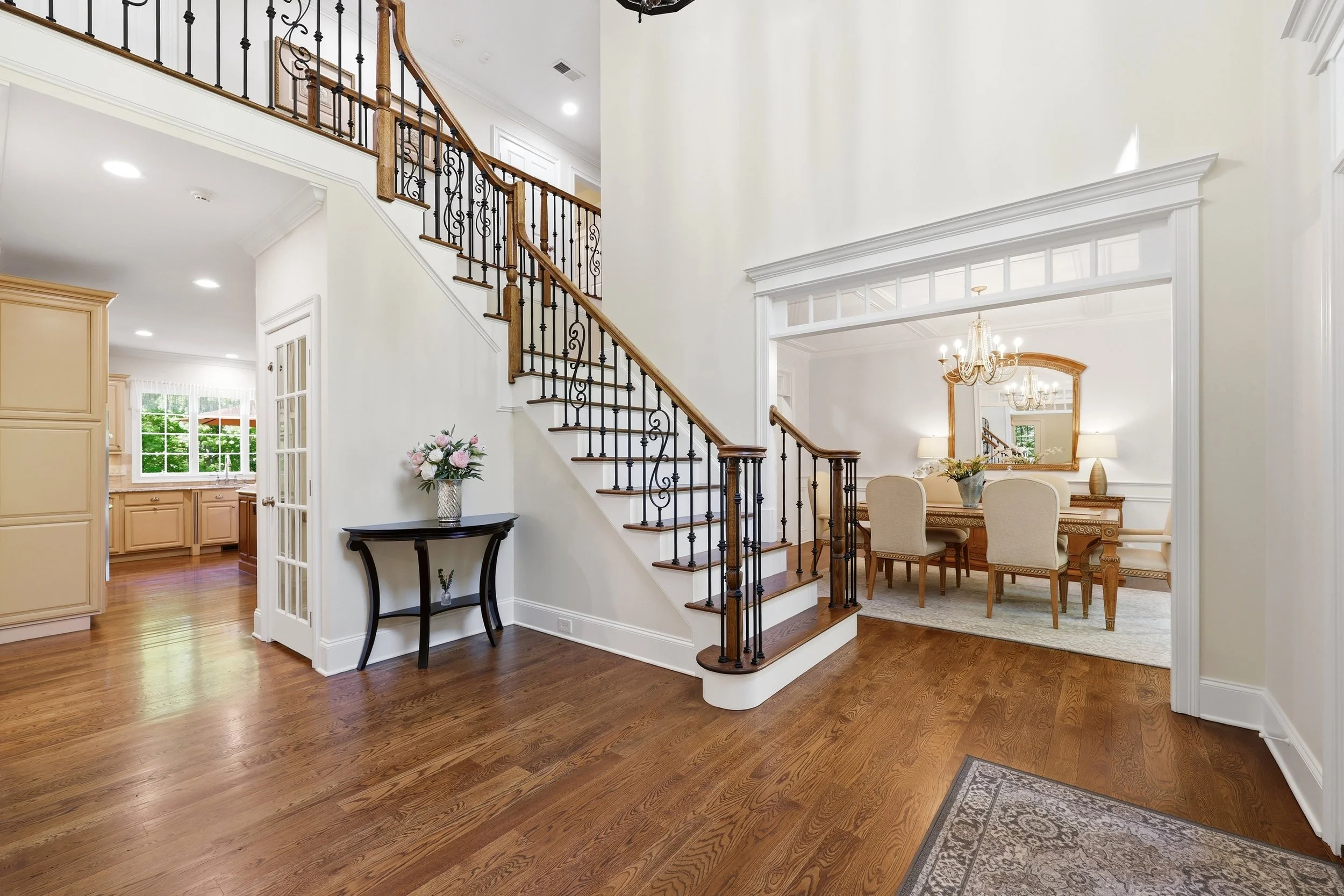 Interior view of a spacious living area with hardwood floors, a staircase with wooden handrails and decorative black iron balusters, a dining area with a chandelier and a mirror, and a kitchen with light-colored cabinets and a window showing green ou