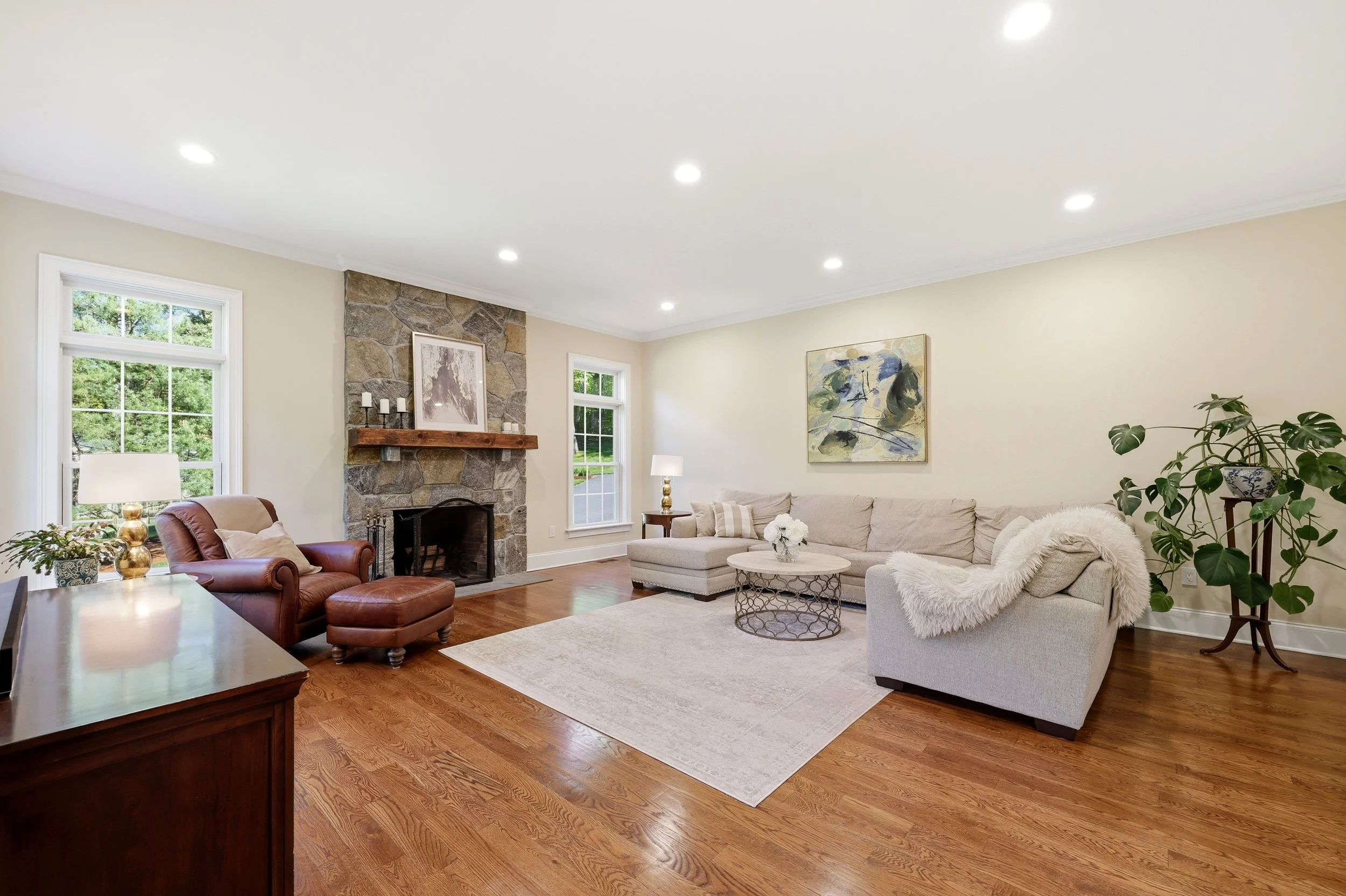 Living room with a stone fireplace, beige walls, hardwood floors, large windows, and a mix of modern and traditional furniture, including a beige sofa, leather armchair, and decorative plants.