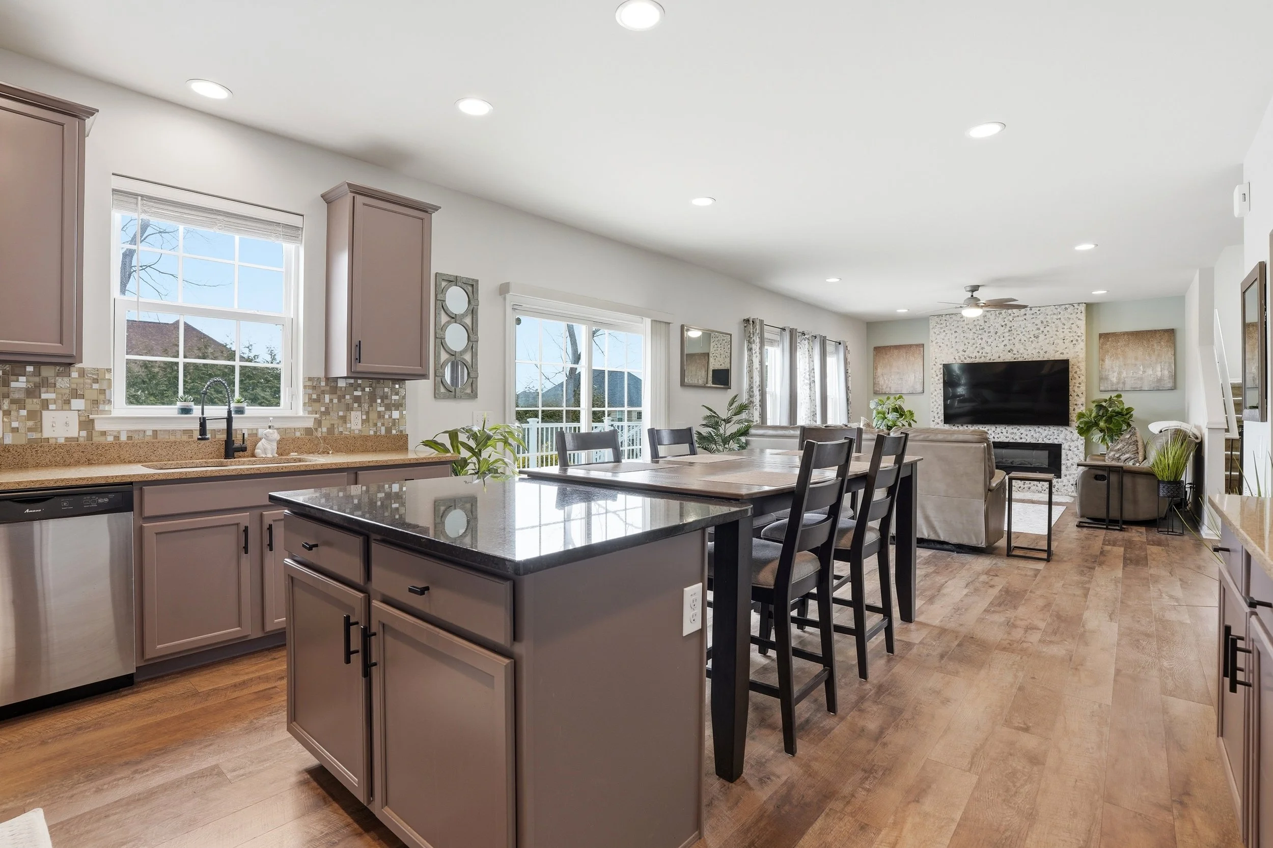 Open-concept kitchen and living room with beige cabinets, granite countertops, a tile backsplash, and a dining area. The living room has a large television on a patterned accent wall and a ceiling fan. Multiple windows let in natural light, and there