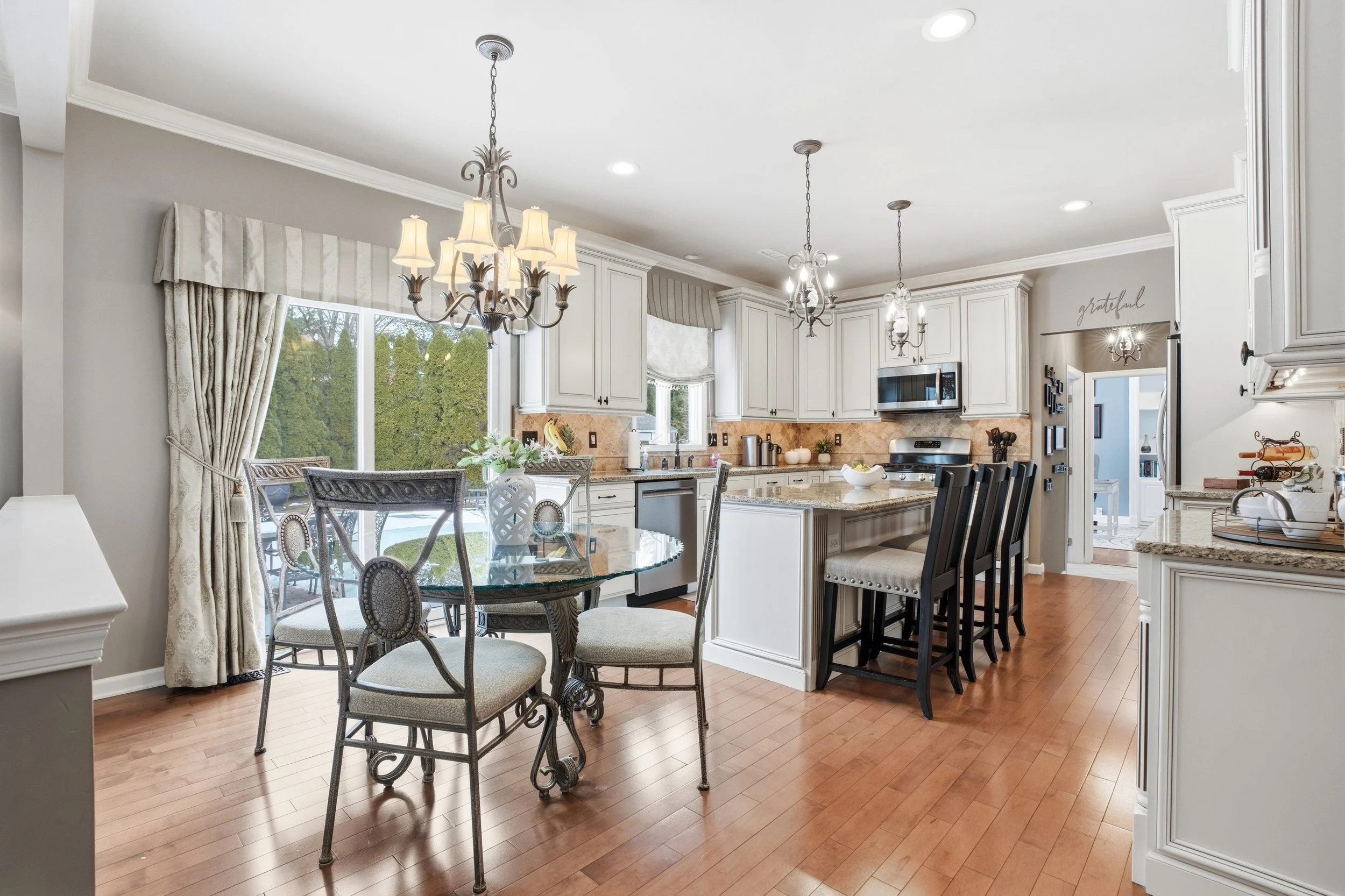 A spacious kitchen and dining area with white cabinetry, granite countertops, and pendant lighting. There is a glass-top dining table with chairs, large window with curtains, and hardwood floors.